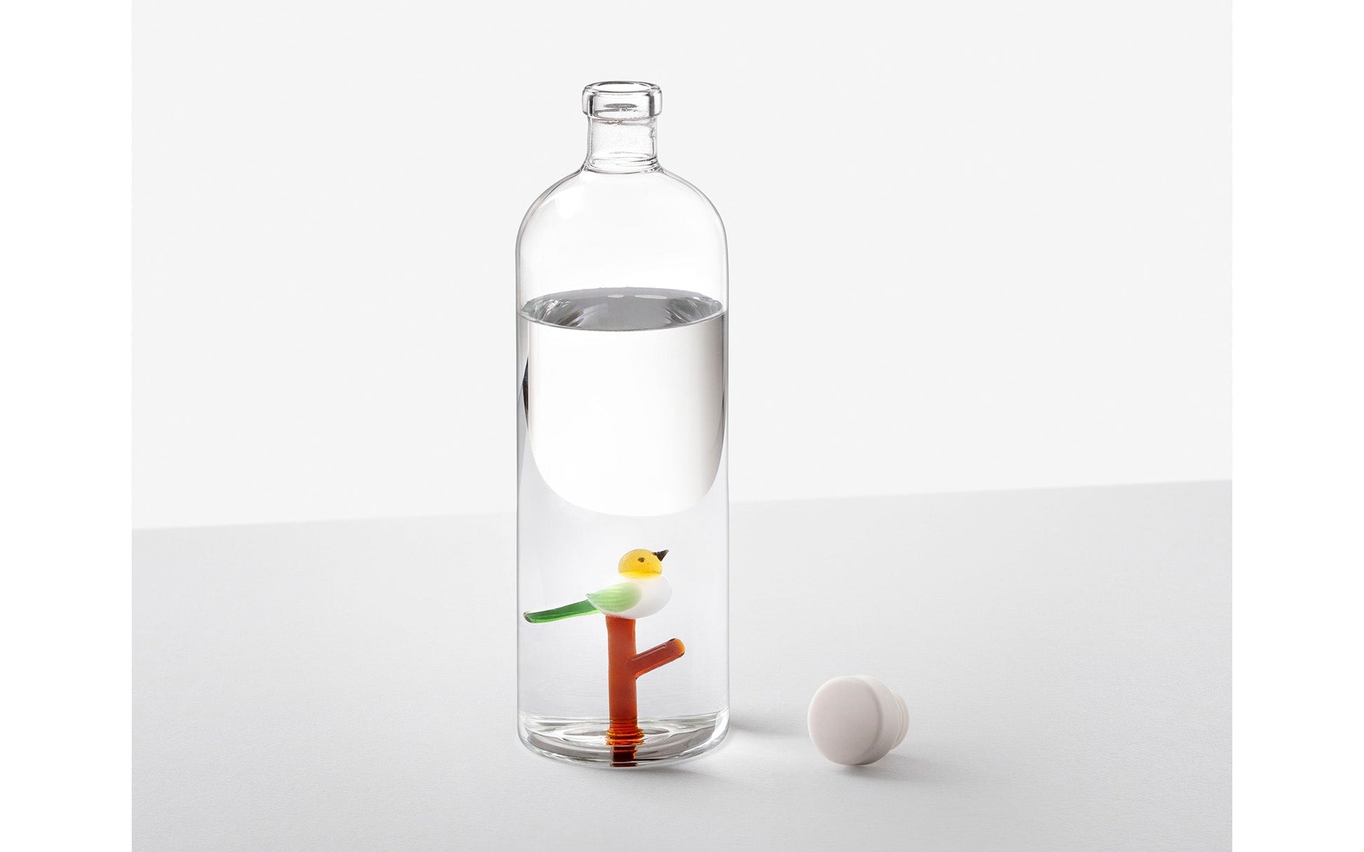 The Ichendorf Milano Animal Farm Bottle by Ichendorf Milano is a unique glass bottle filled with water, showcasing a small decorative bird on an orange branch inside. Its white cap sits beside it, all set against a plain white background.