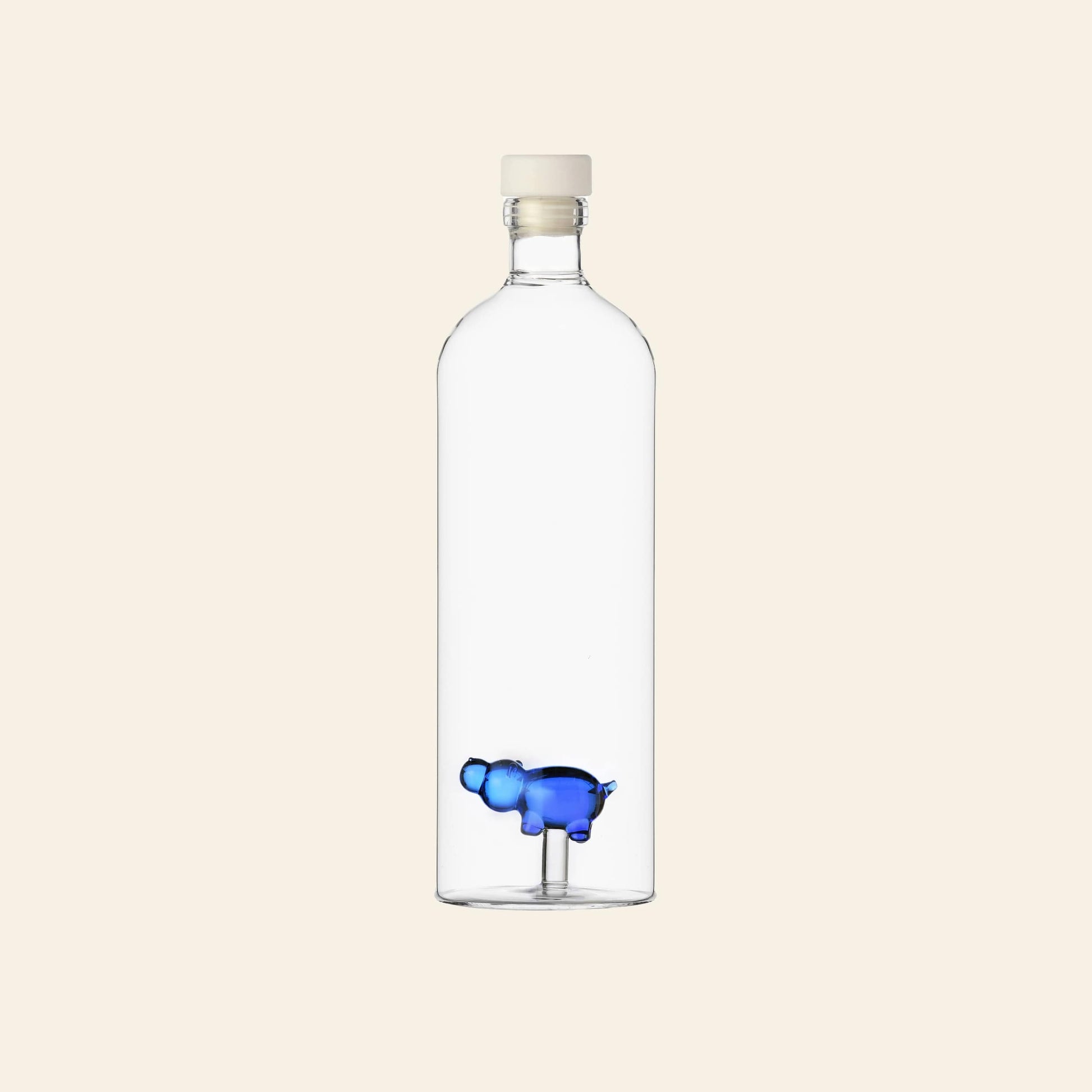 The Ichendorf Milano Animal Farm Bottle by Ichendorf Milano features a unique glass design with a white cap and a small blue hippopotamus figurine near the bottom, set against a plain, light-colored background.