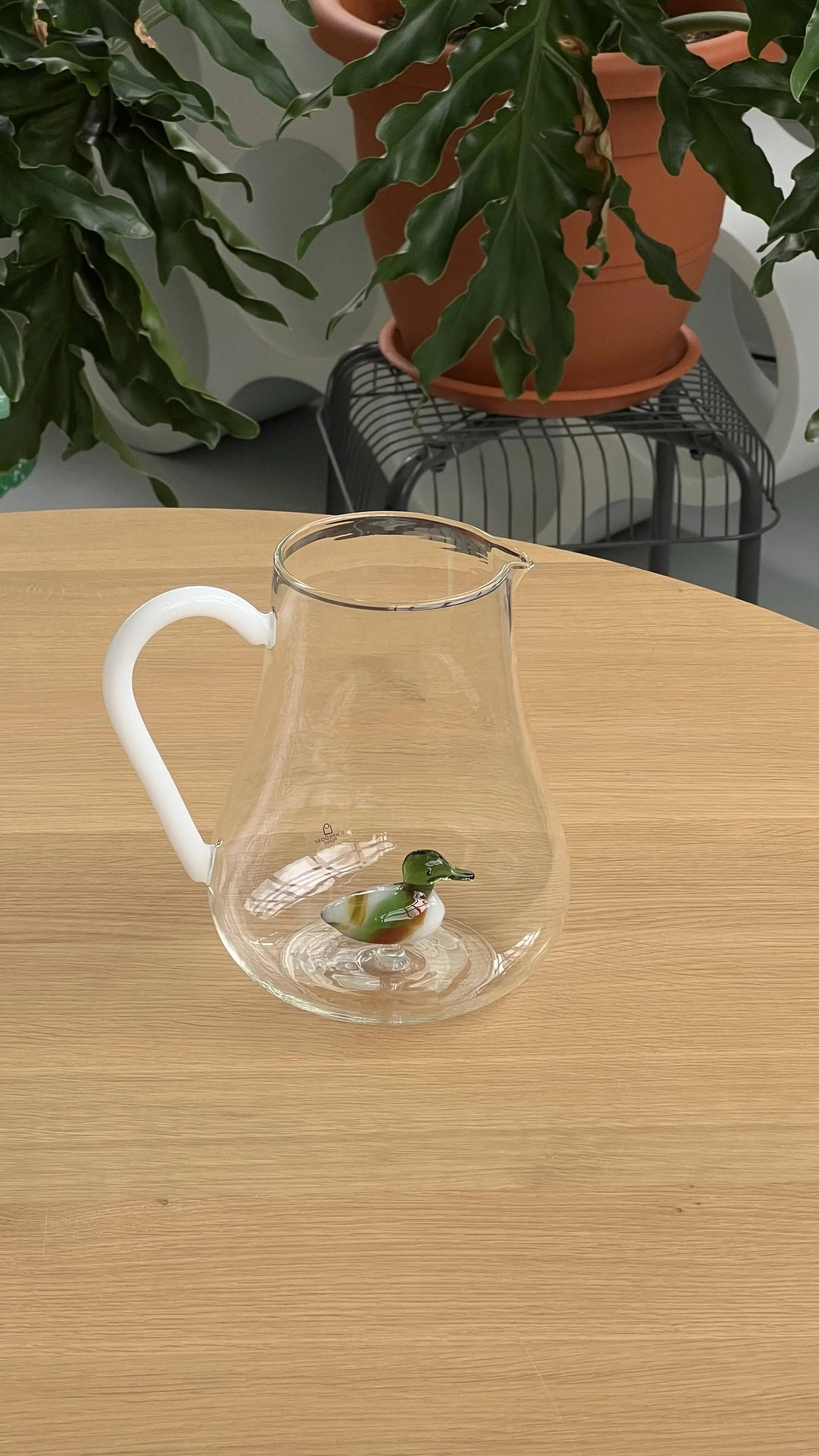 The Ichendorf Milano Animal Farm Pitcher - Duck by Stacked Store, with a clear glass body and white handle, sits on a wooden table. Inside is a decorative glass duck, with green potted plants in the background.