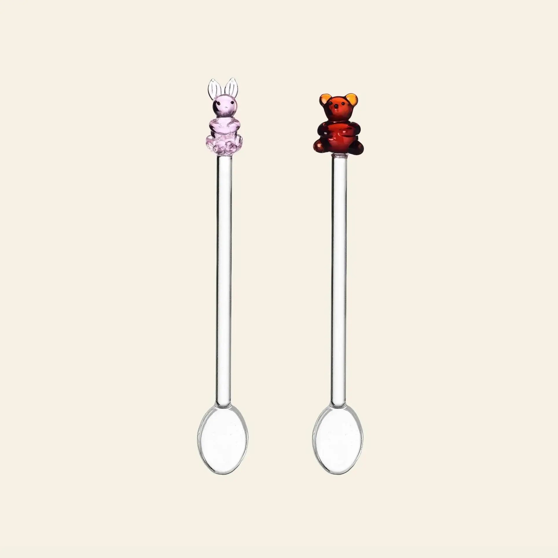 The Ichendorf Milano Animal Farm Spoons, Set of 2, features clear glass spoons with decorative handles—one topped with a pink bunny, the other with a brown bear—displayed on a plain light background.