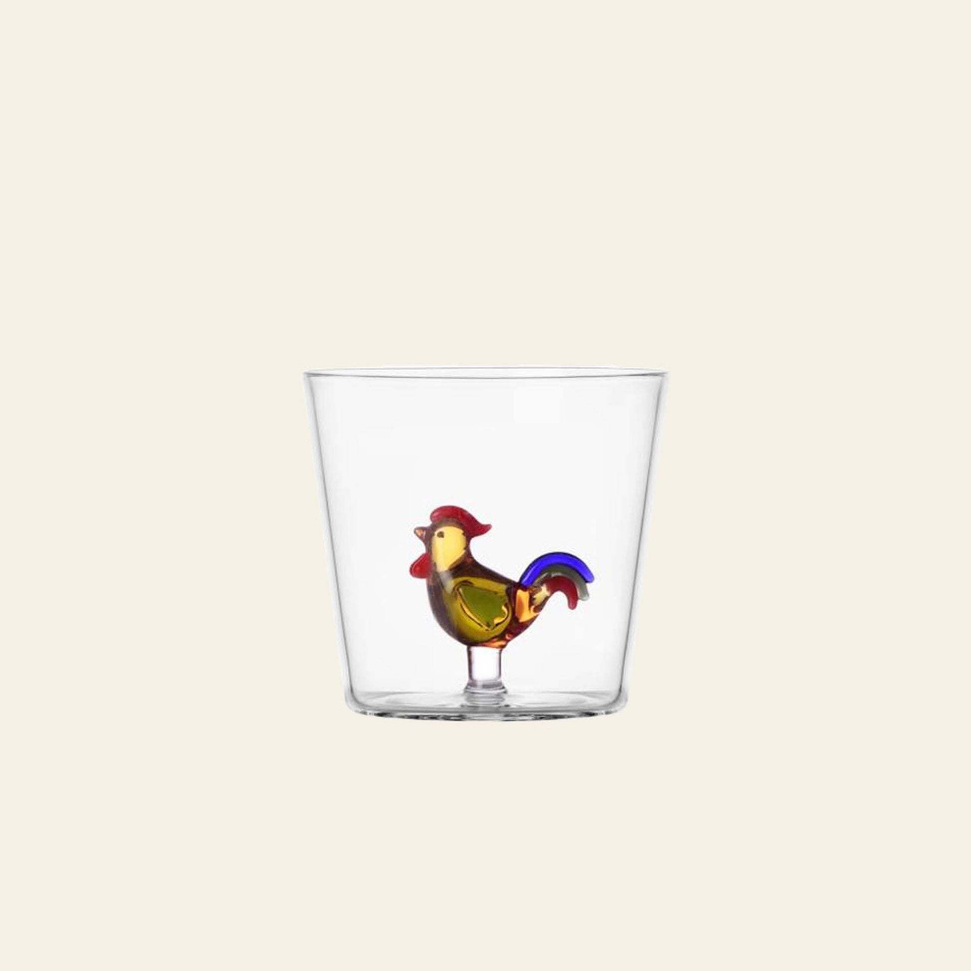 The Ichendorf Milano Animal Farm Tumbler by Ichendorf Milano is a clear borosilicate glass tumbler featuring a colorful rooster figurine with red, yellow, and blue accents at the base, set against a plain off-white background.