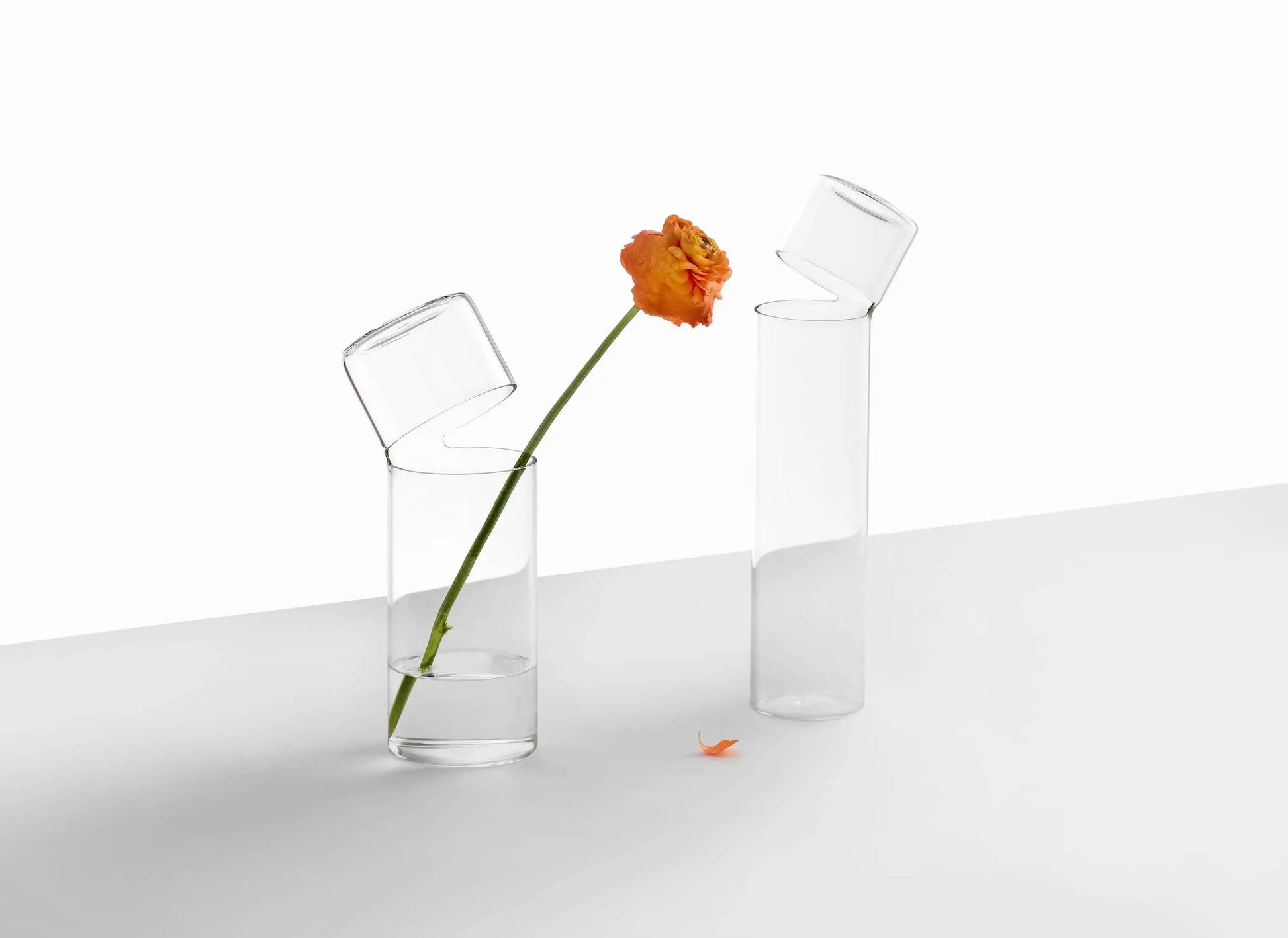 Two medium Ichendorf Milano Attesa Flower Vases sit on a white surface. One holds an orange flower with a bent stem and scattered petals, while the other is empty. Both feature slanted openings that highlight their handcrafted charm.