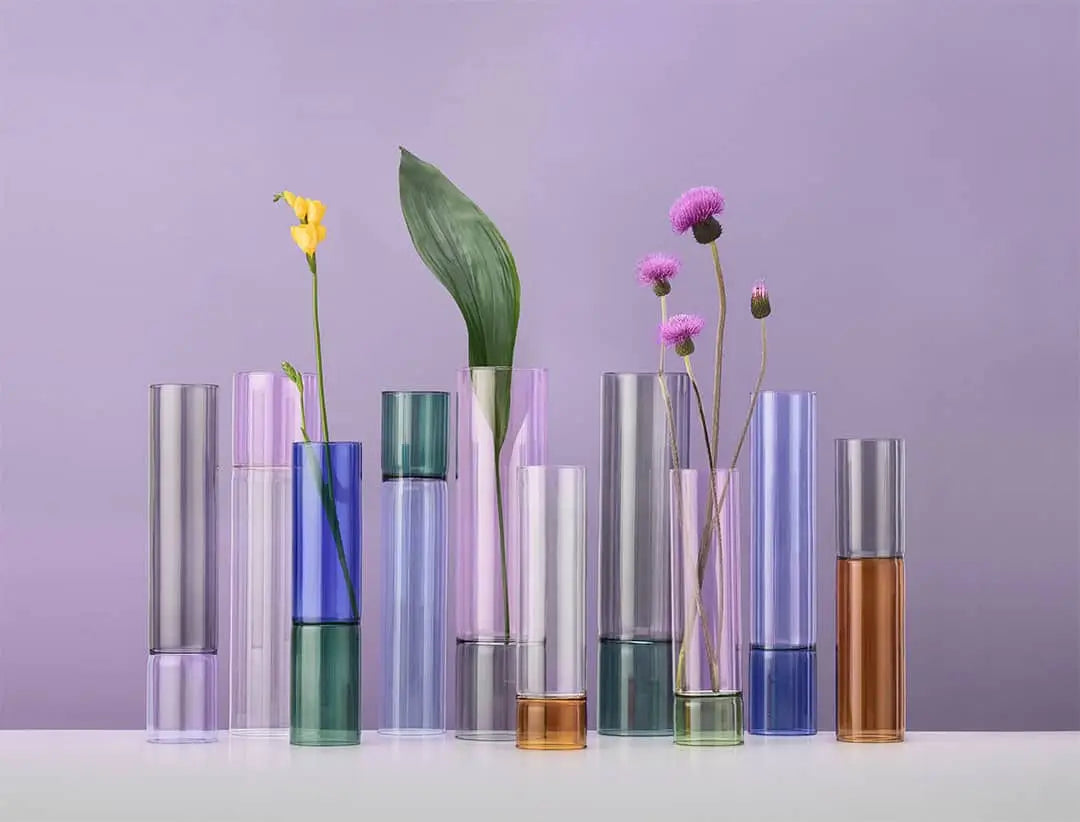 The Ichendorf Milano Bamboo Groove Vases (30cm) by Ichendorf Milano—tall, cylindrical, and grooved in various colors—hold single flower stems and leaves on a white surface with a light purple background.