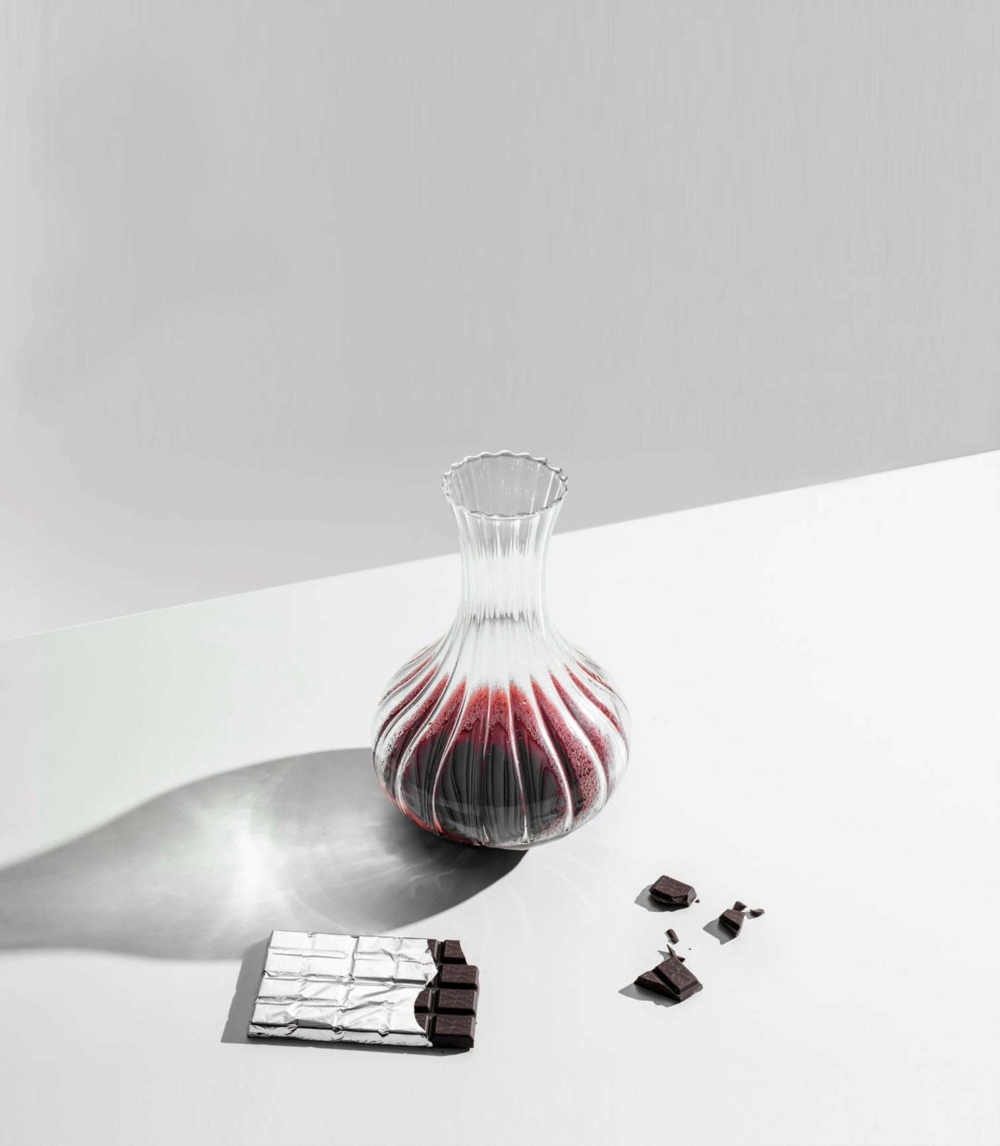The Ichendorf Milano Bistrot Decanter by Ichendorf Milano, filled with red wine, rests on a white surface. Nearby are pieces of dark chocolate—some broken—and a partially unwrapped silver chocolate bar.