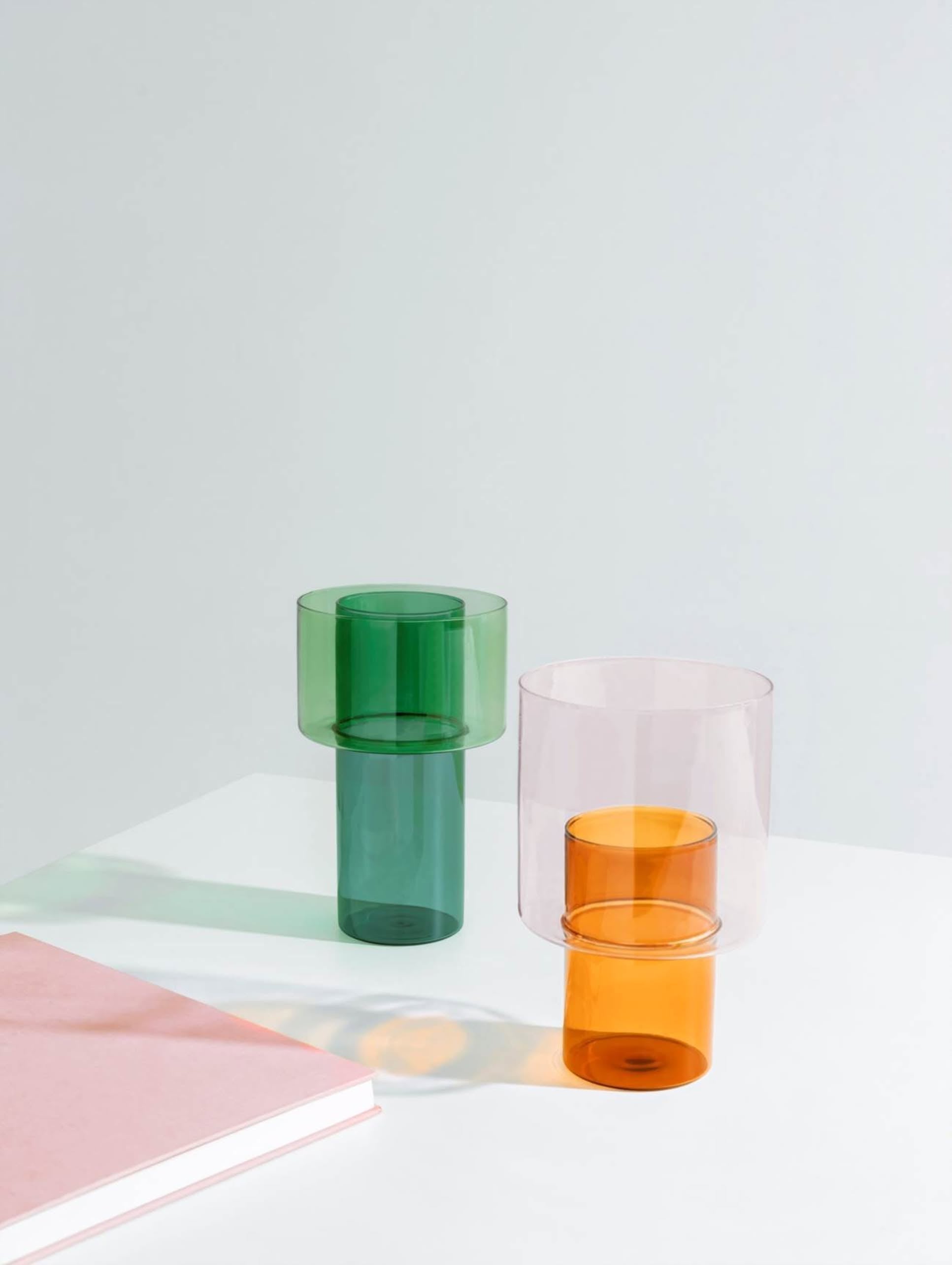 The Ichendorf Milano Boreale Vase B by Ichendorf Milano stands on a white surface, casting colorful shadows. A closed pink book lies in the bottom left corner, set against a simple light grey background.