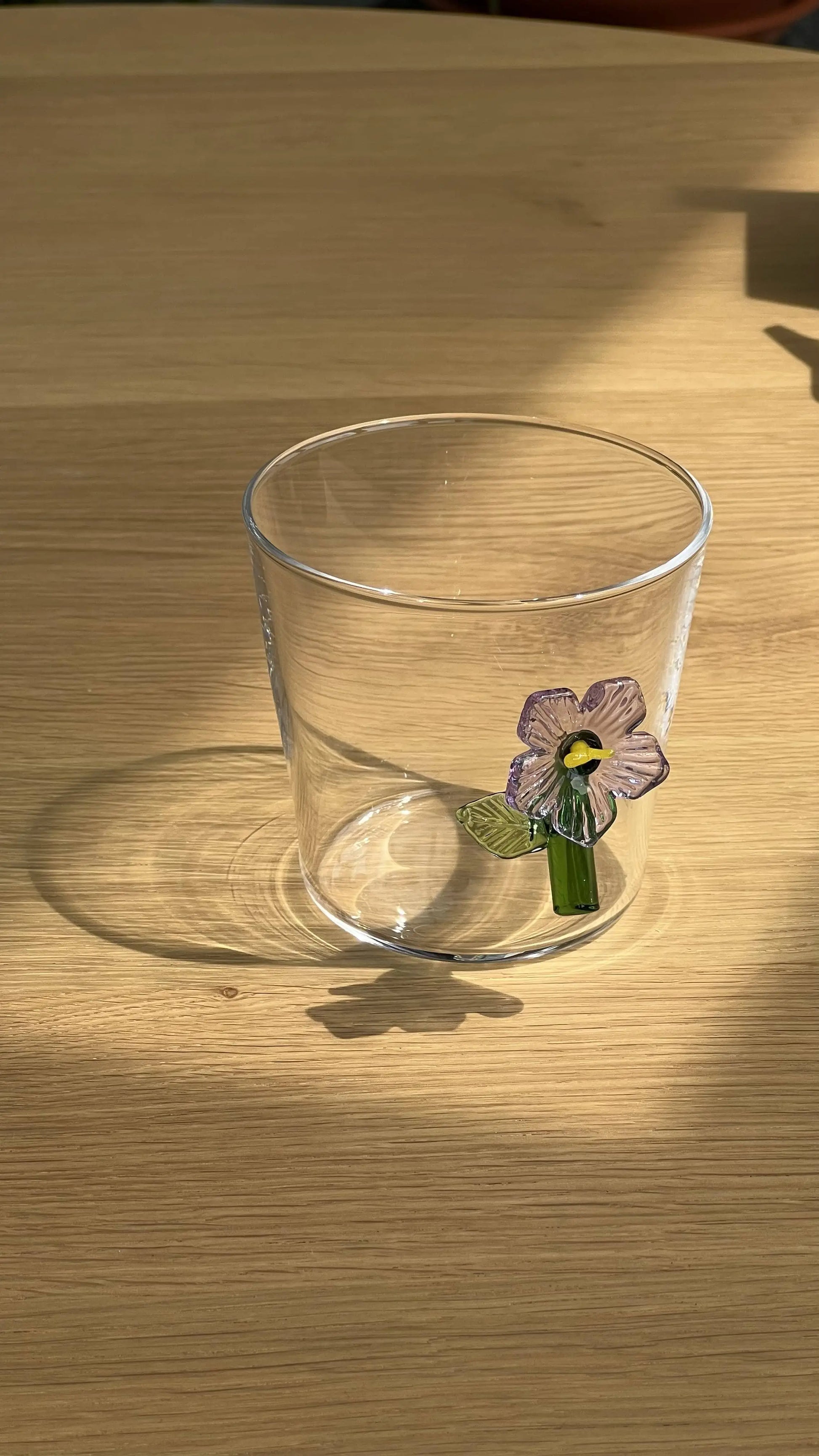 The Ichendorf Milano Botanica Tumbler - Lilac, featuring a delicate purple flower and green leaf motif, sits on a wooden surface, casting a clear shadow and adding nature-inspired charm to your space.