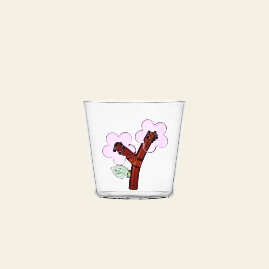 The Ichendorf Milano Botanica Tumbler - Peach Blossom is a clear glass tumbler featuring two cartoonish brown tree branches with smiling faces, pink flowers, and green leaves on a beige background. Brand: Ichendorf Milano.