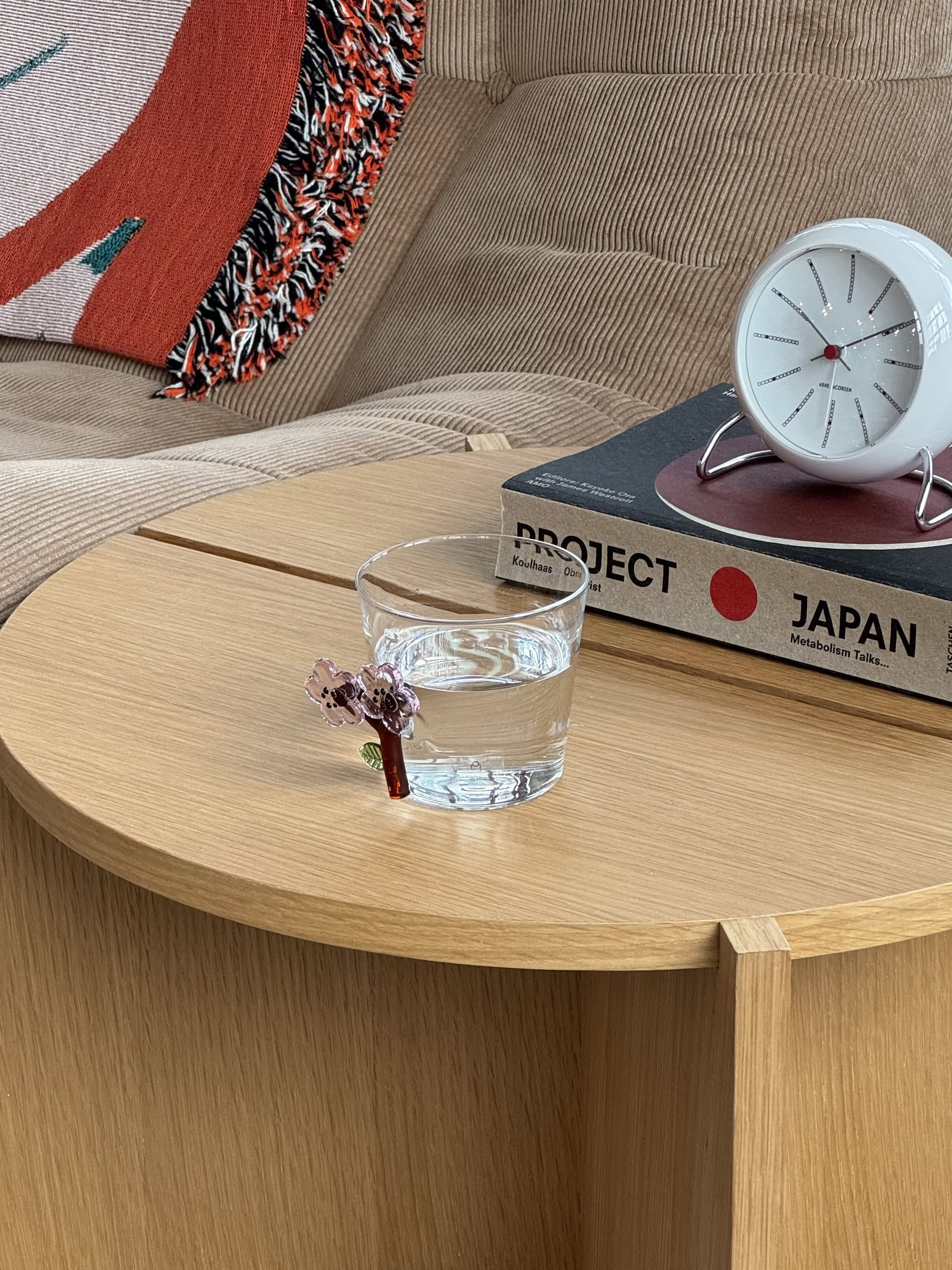 A glass of water in the Ichendorf Milano Botanica Tumbler - Peach Blossom sits on a wooden table with PROJECT JAPAN, a white clock, beige sofa, and colorful cushion, creating a serene scene inspired by Ichendorf Milano’s floral design.