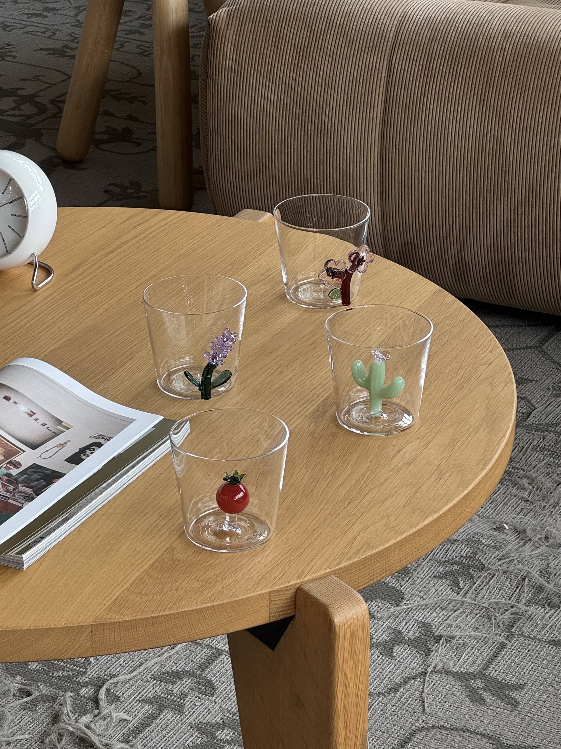 An Ichendorf Milano Botanica Tumbler - Peach Blossom sits on a round wooden table beside a clock, an open magazine, and a striped sofa in a cozy living room.