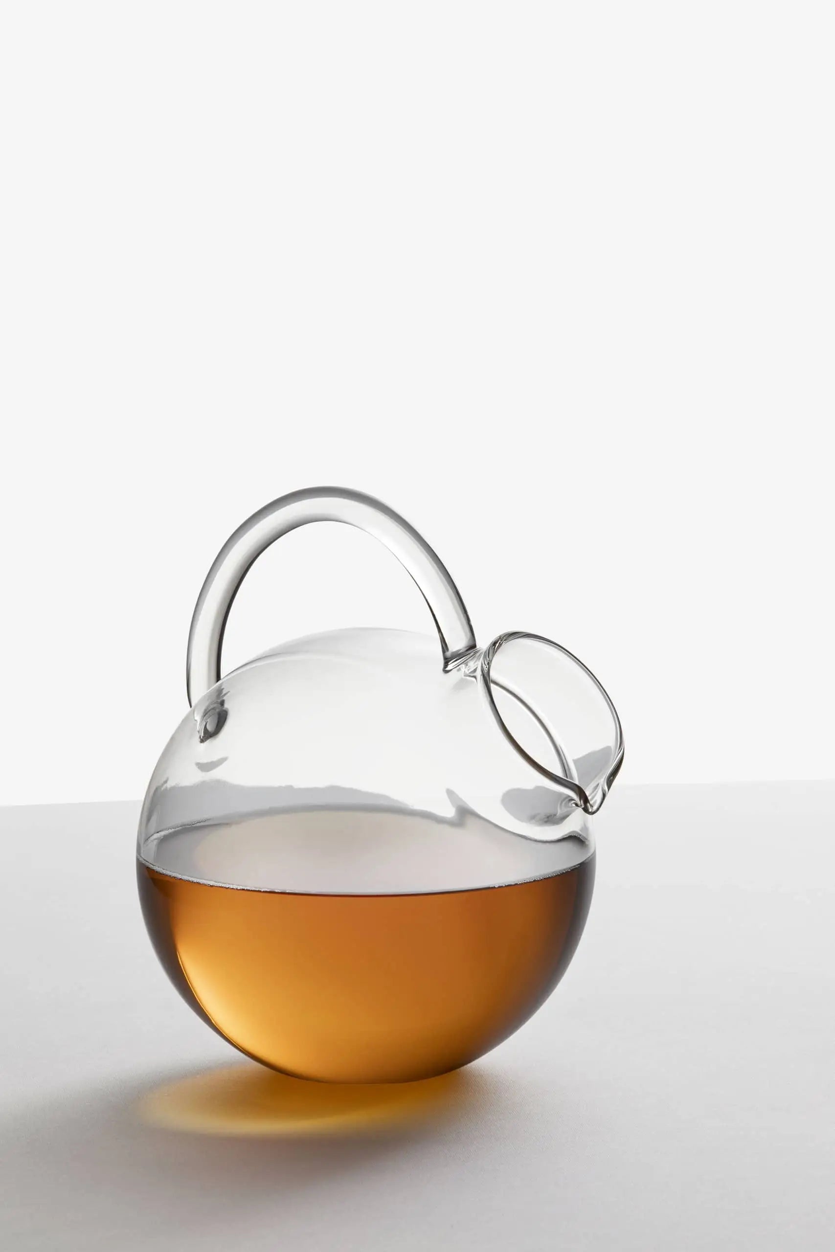 The Ichendorf Milano Bouquet Round Jug by Ichendorf Milano, crafted in clear glass with a round body and arched handle, sits partially filled with amber liquid on a white surface against a light background.