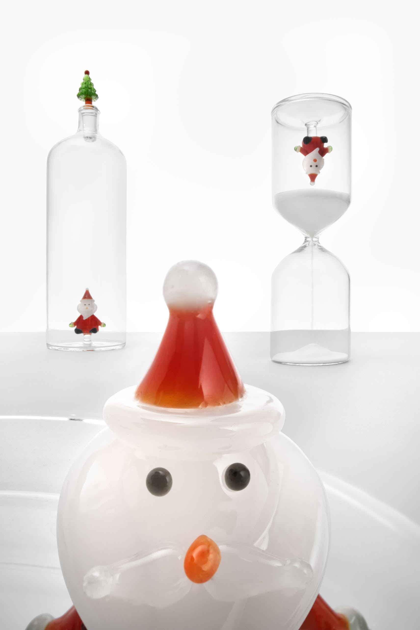 A glass Santa figurine stands in front. Behind it, the Ichendorf Milano Christmas Tales Bottle by Ichendorf Milano features a tiny Christmas tree on top and another Santa upside down in an hourglass, all set against a white background.