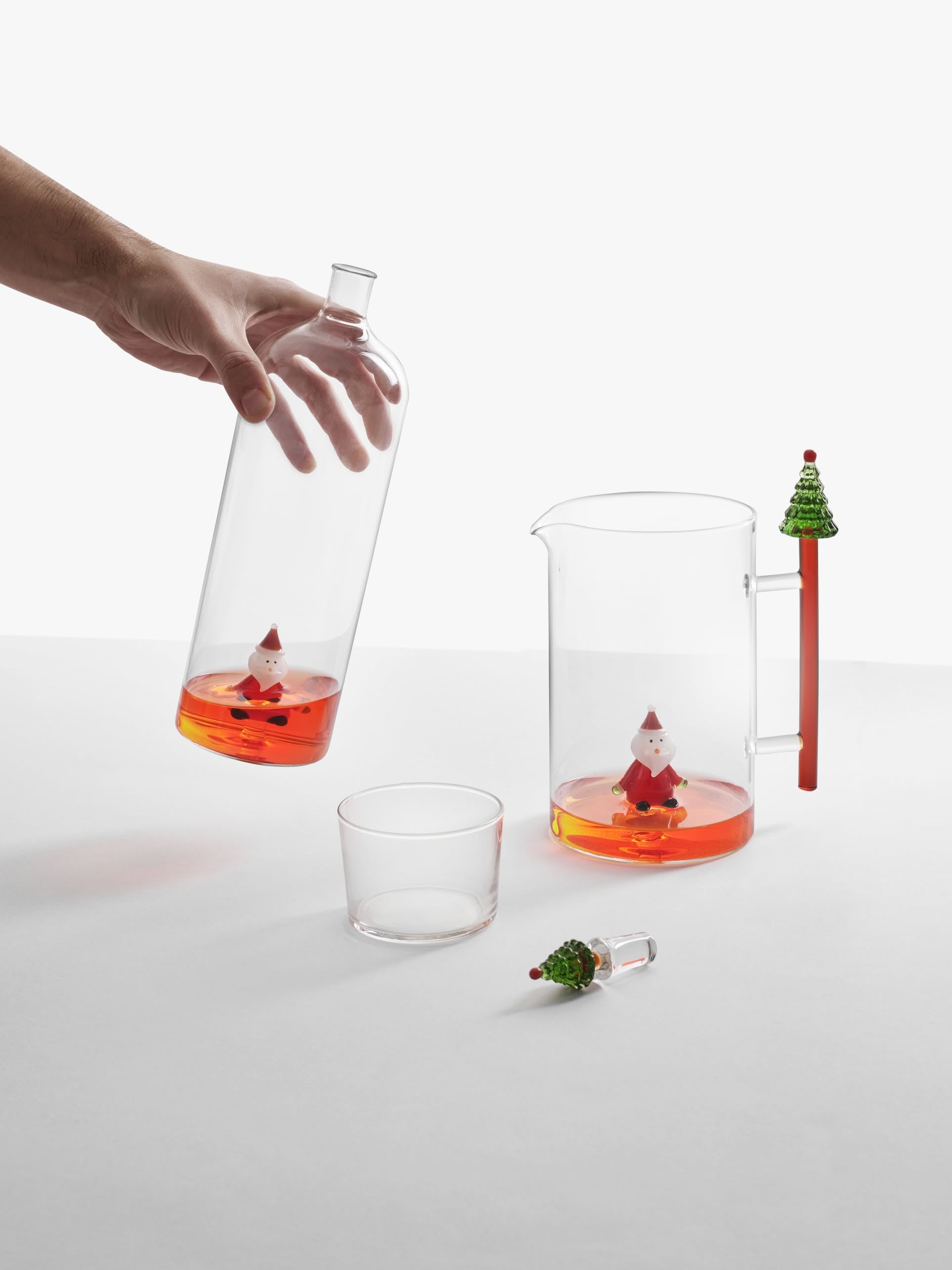A hand pours orange liquid from the Ichendorf Milano Christmas Tales Bottle by Ichendorf Milano into a glass. On the white table are a clear pitcher with more orange liquid, the festive bottle inside, a tree-topped stirrer, and a glass.