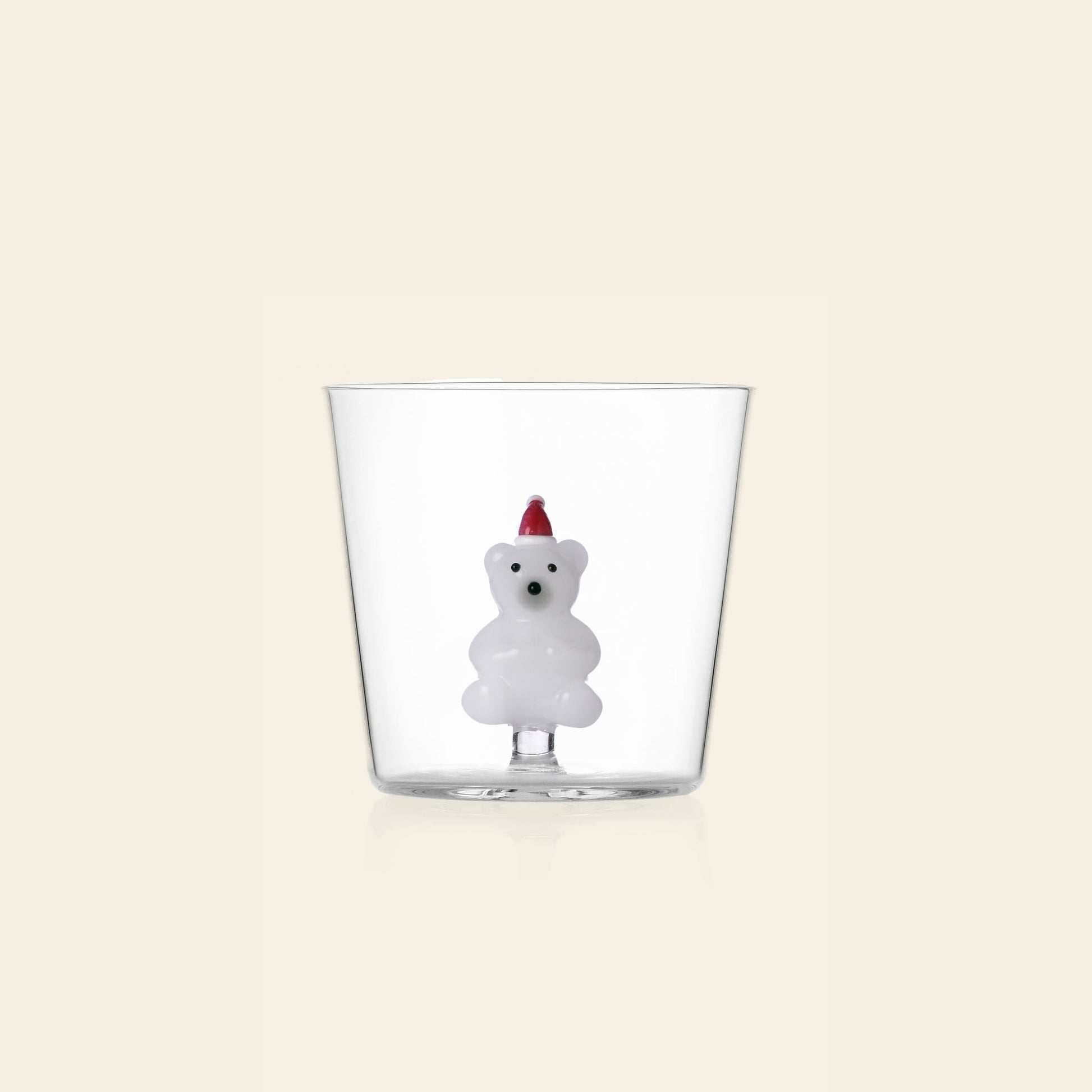 The Ichendorf Milano Christmas Tales Tumbler by Ichendorf Milano features a clear glass cup with a small white bear figurine in a red party hat attached inside—ideal as festive glassware or Christmas decor, shown on a light beige background.