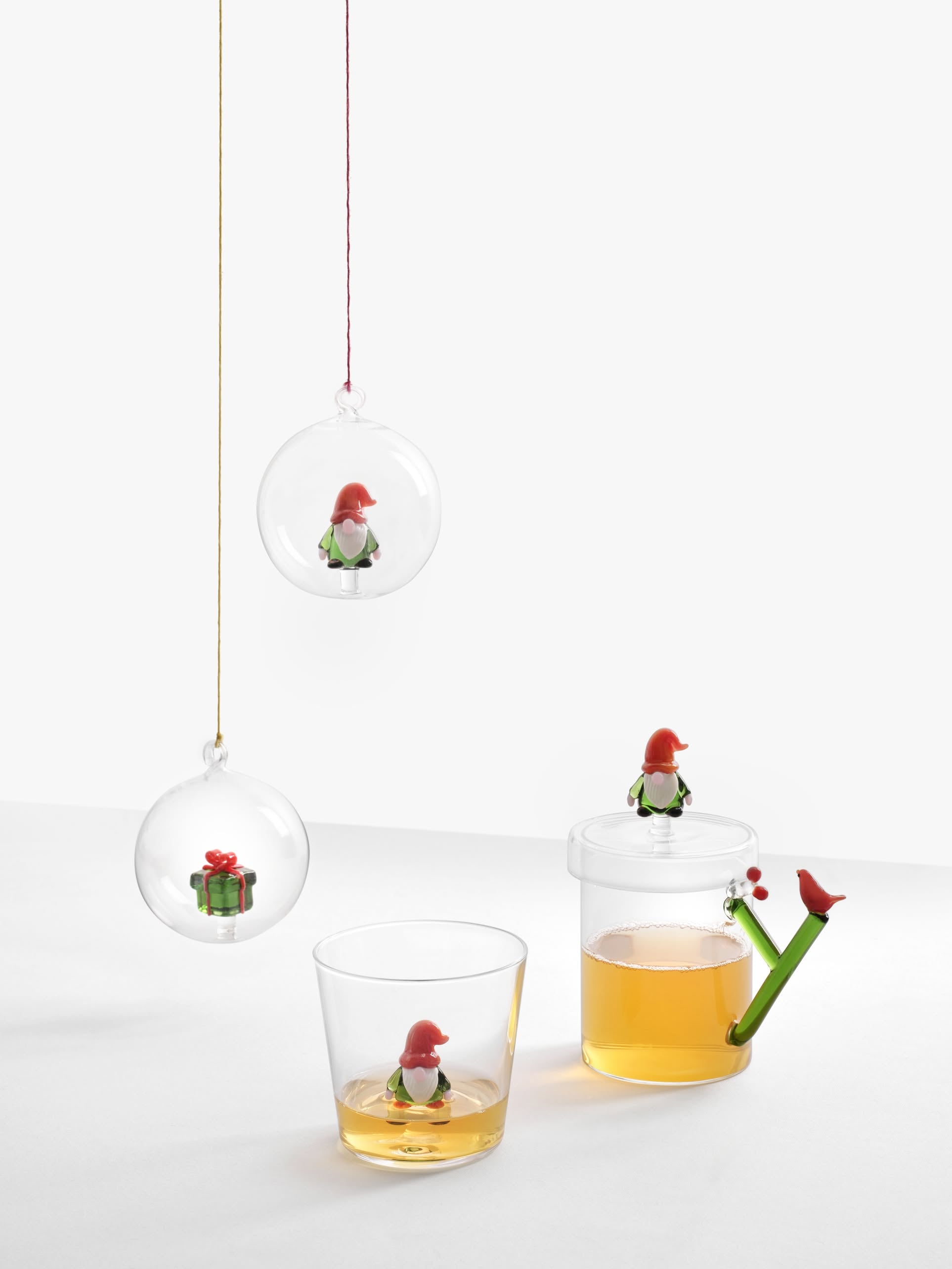 Two clear ornaments hang, one holding a gnome and the other a gift. Below, a gnome sits on Ichendorf Milano Christmas Tales glassware—a glass and festive tumbler—each filled with amber liquid, perfect for enjoying your favorite holiday drink.