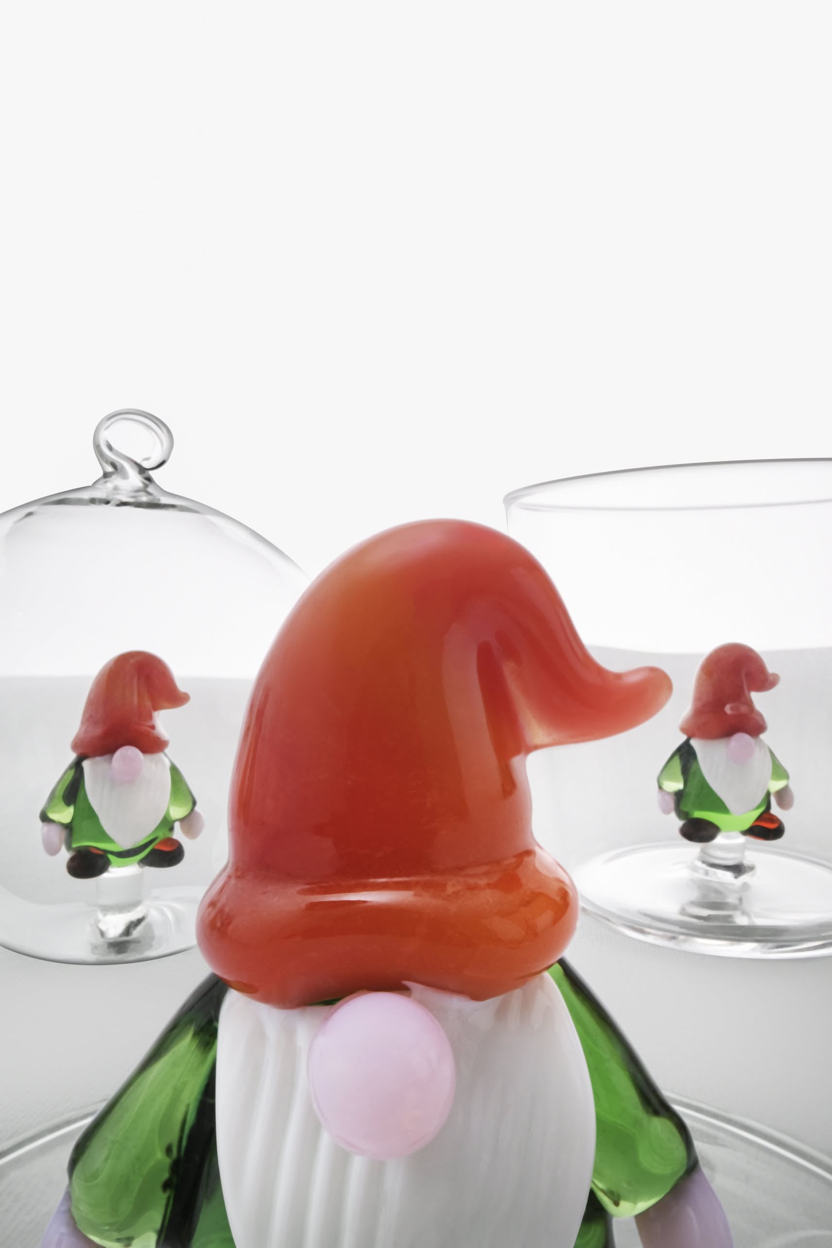 A close-up of the Ichendorf Milano Christmas Tales Tumbler, a colorful glass gnome figurine by Ichendorf Milano with an orange hat and green coat, perfect for holiday gifting, with two smaller gnome tumblers in glass containers in the background.