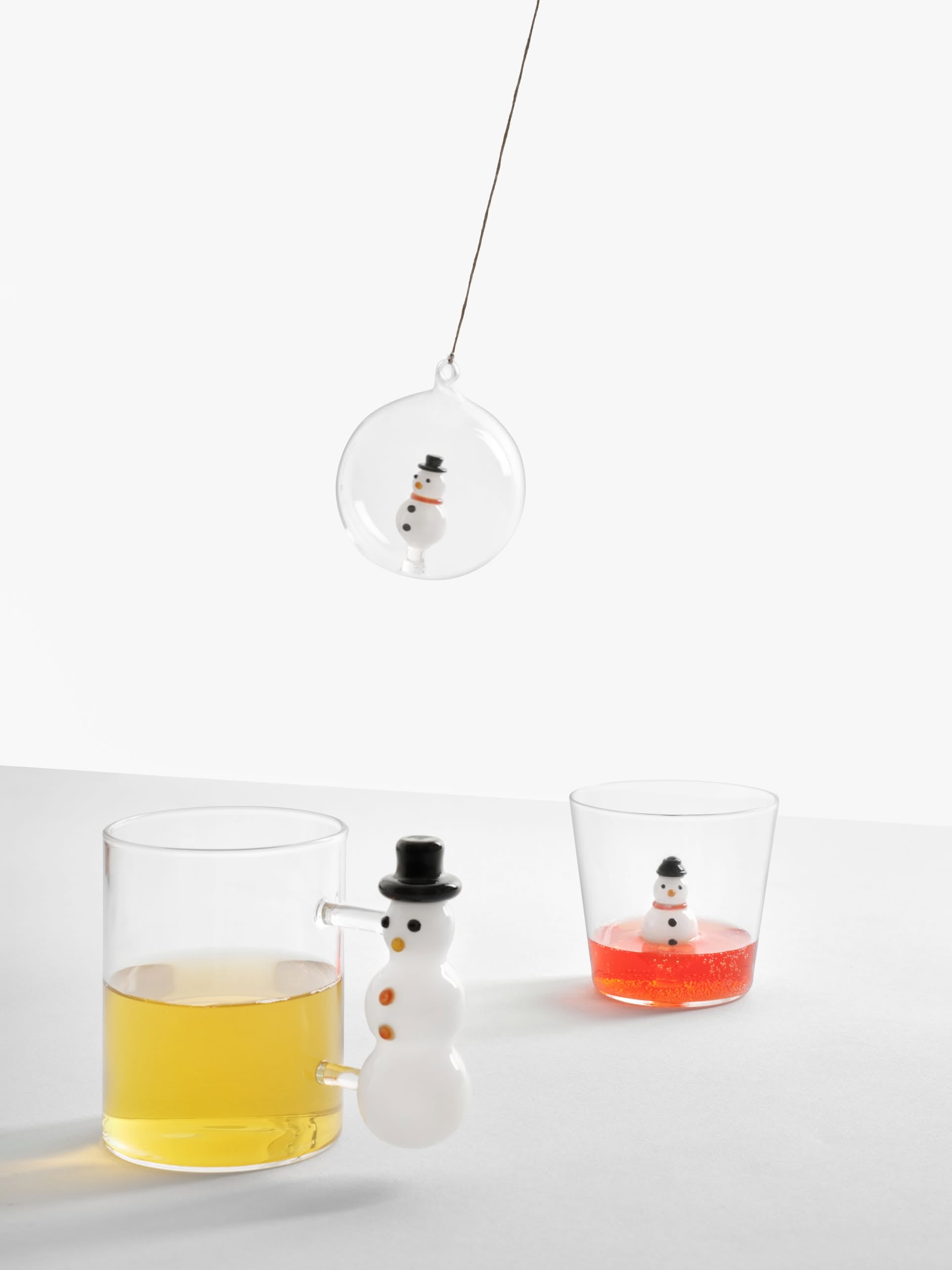 The Ichendorf Milano Christmas Tales Tumbler by Ichendorf Milano features three glass snowmen: one inside a clear ornament, one on the rim of a festive tumbler with yellow liquid, and one in a glass of red—perfect for unique Christmas glassware or gifting.