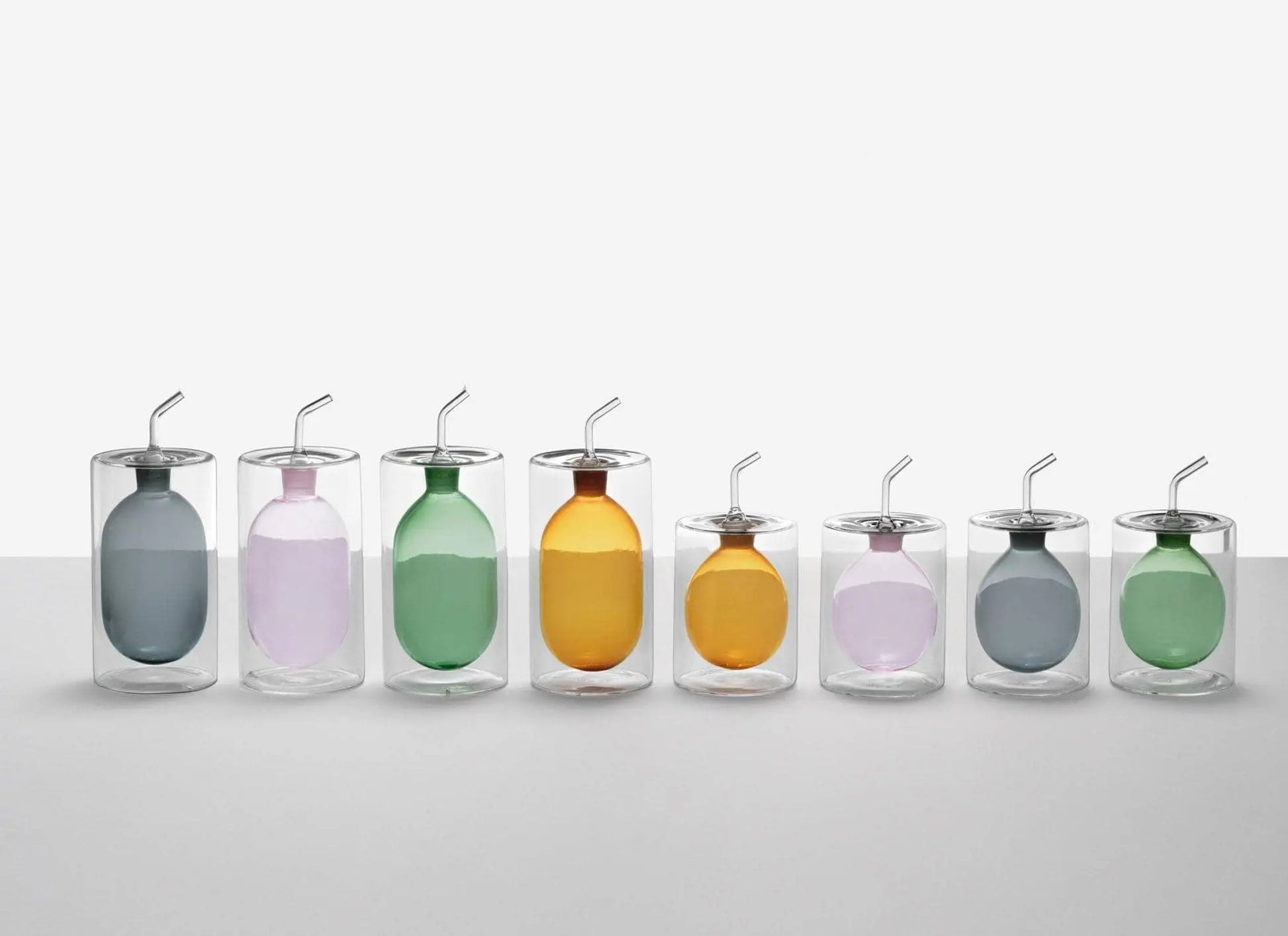 Eight transparent glasses with metal straws and colorful balloons are arranged in a row against a plain white background, echoing the modern, sleek design of the Ichendorf Milano Cilindro Oil Bottle by Ichendorf Milano.
