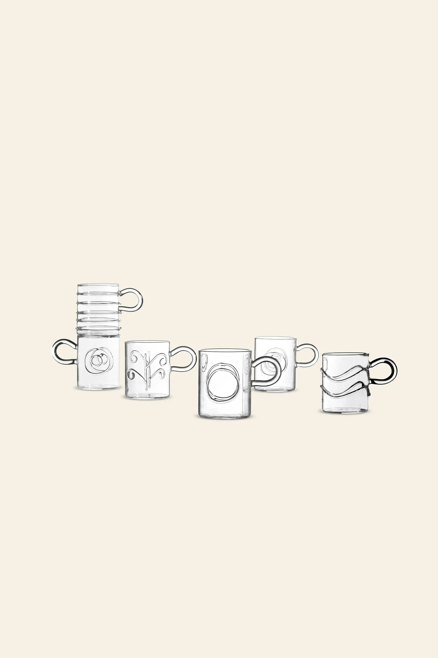 Six Ichendorf Milano Deco Coffee Cups, each with a unique shape and artistic handle, are arranged in a row on a plain beige background. Crafted from borosilicate glass, these minimalist mugs showcase distinct etched designs. Brand: Ichendorf Milano.