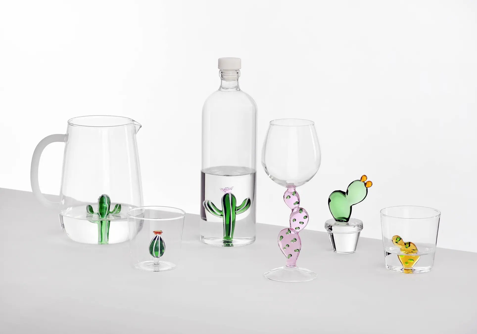 The Ichendorf Milano Desert Plants Pitcher, part of the handcrafted glassware collection by Ichendorf Milano, features a colorful cactus motif and is displayed with matching bottle, wine glass, cocktail glass, and tumblers on a white surface.