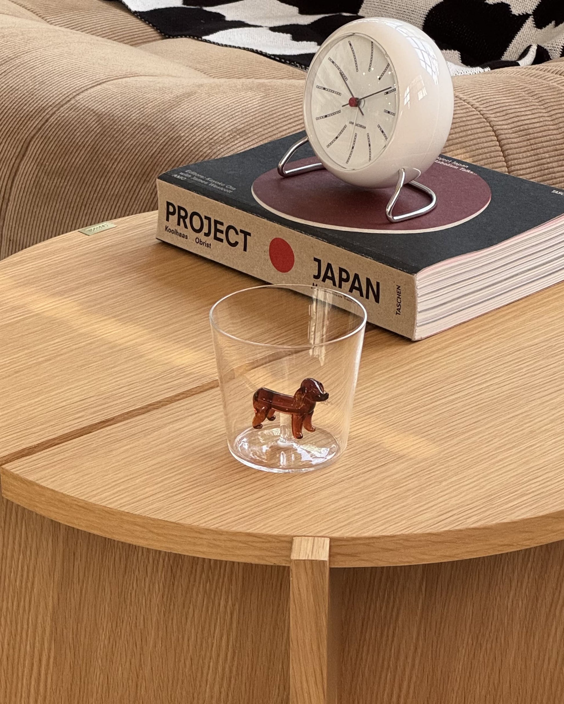 An Ichendorf Milano Dogs Tumbler - Dachshund sits on a wooden table beside a book titled Project Japan and a white clock, with a beige couch and patterned blanket in the background.