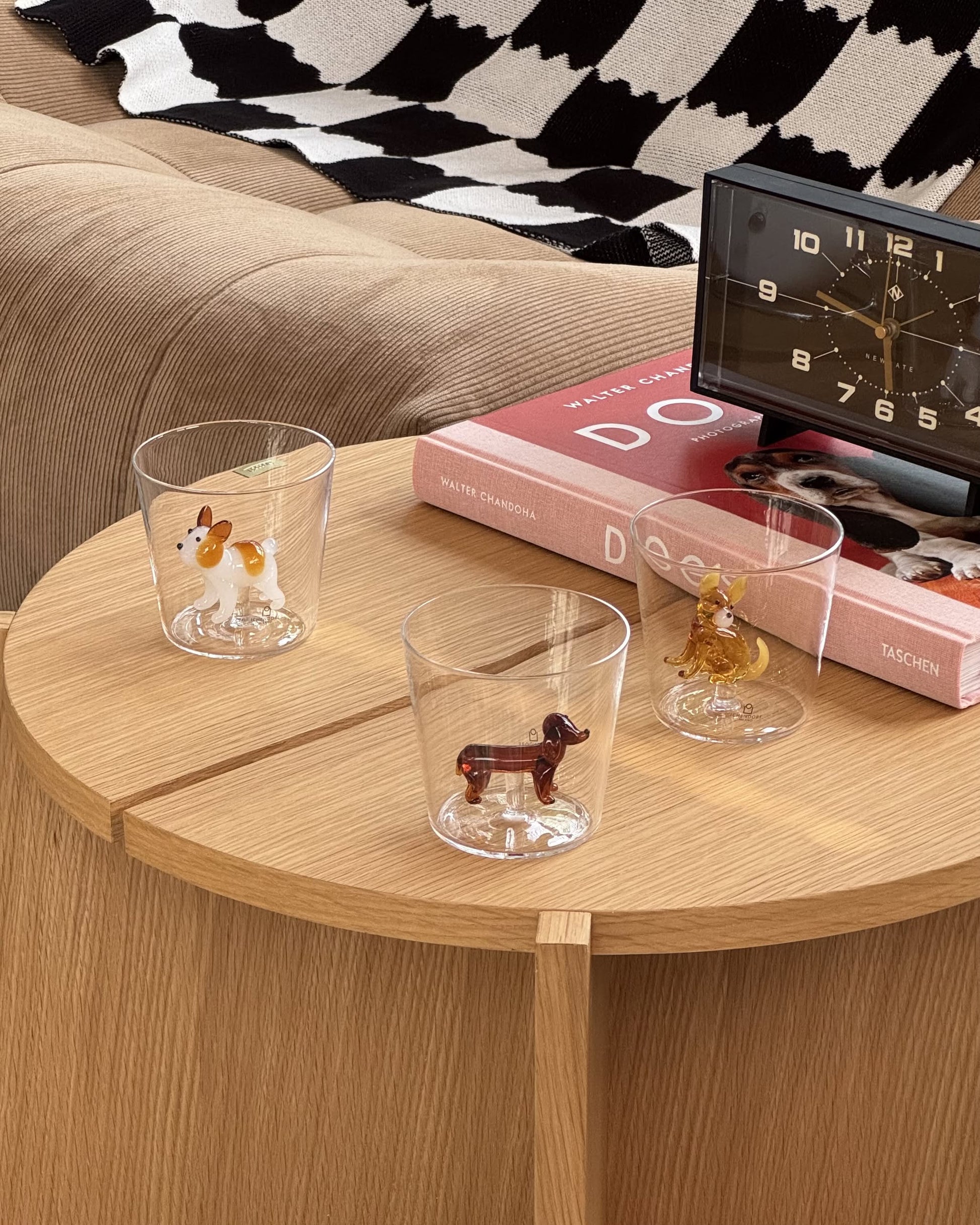 Three clear Ichendorf Milano Dogs Tumblers—featuring dachshund figurines—rest on a light wooden table near two stacked books, a black modern clock, and a beige sofa with a black-and-white throw blanket.