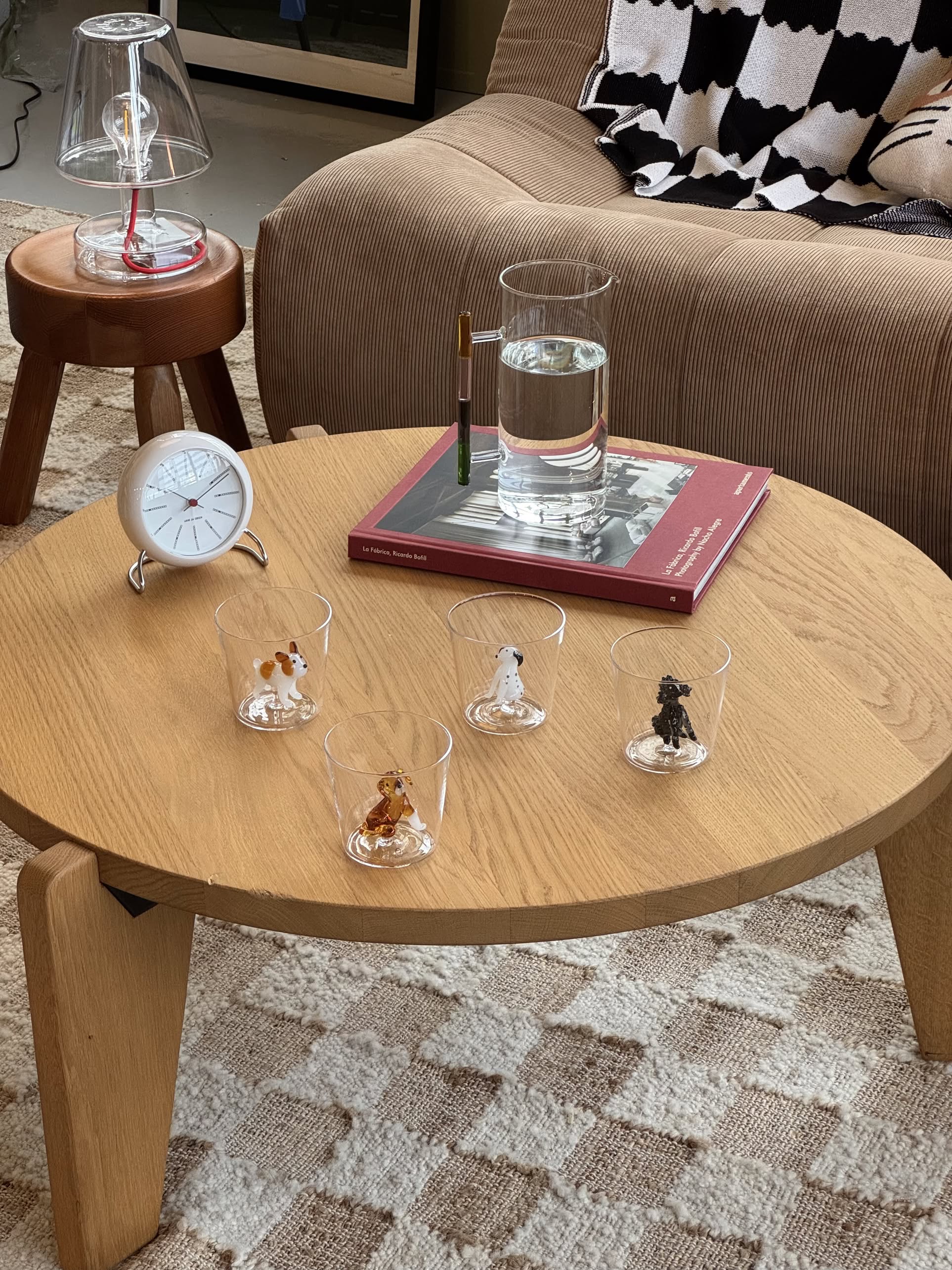 A round wooden coffee table displays five Ichendorf Milano Dogs Tumblers – Dalmatian by Ichendorf Milano, each featuring a Dalmatian figurine. A glass of water, book, clock, pen, tan chair, and checkered rug add to the cozy setting.