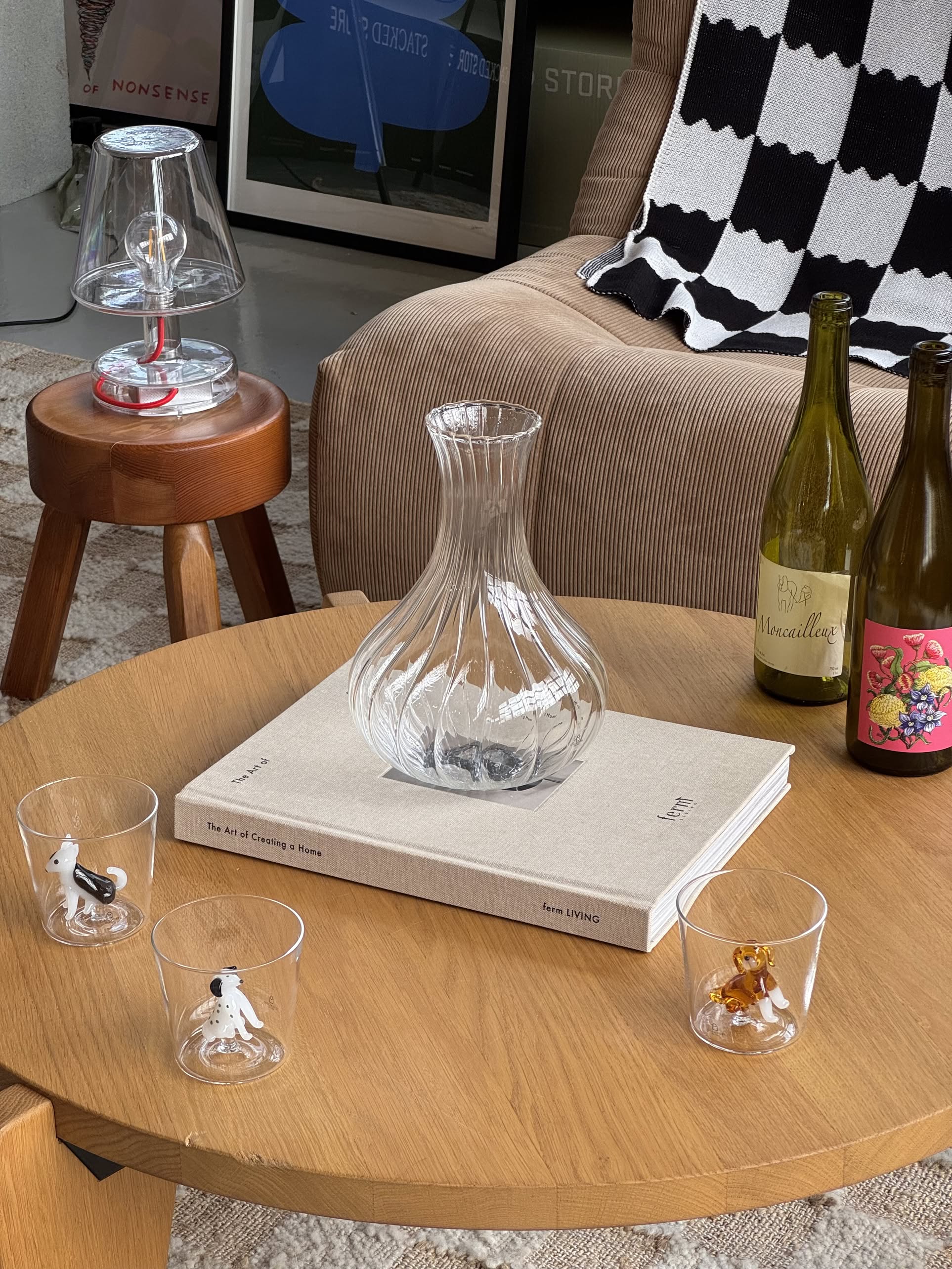 A clear glass carafe rests on a beige book atop a wooden coffee table, surrounded by Ichendorf Milano Dogs Tumblers – Dalmatian, artisan glassware featuring dog figurines, with two wine bottles nearby. A striped blanket and lamp are in the background.