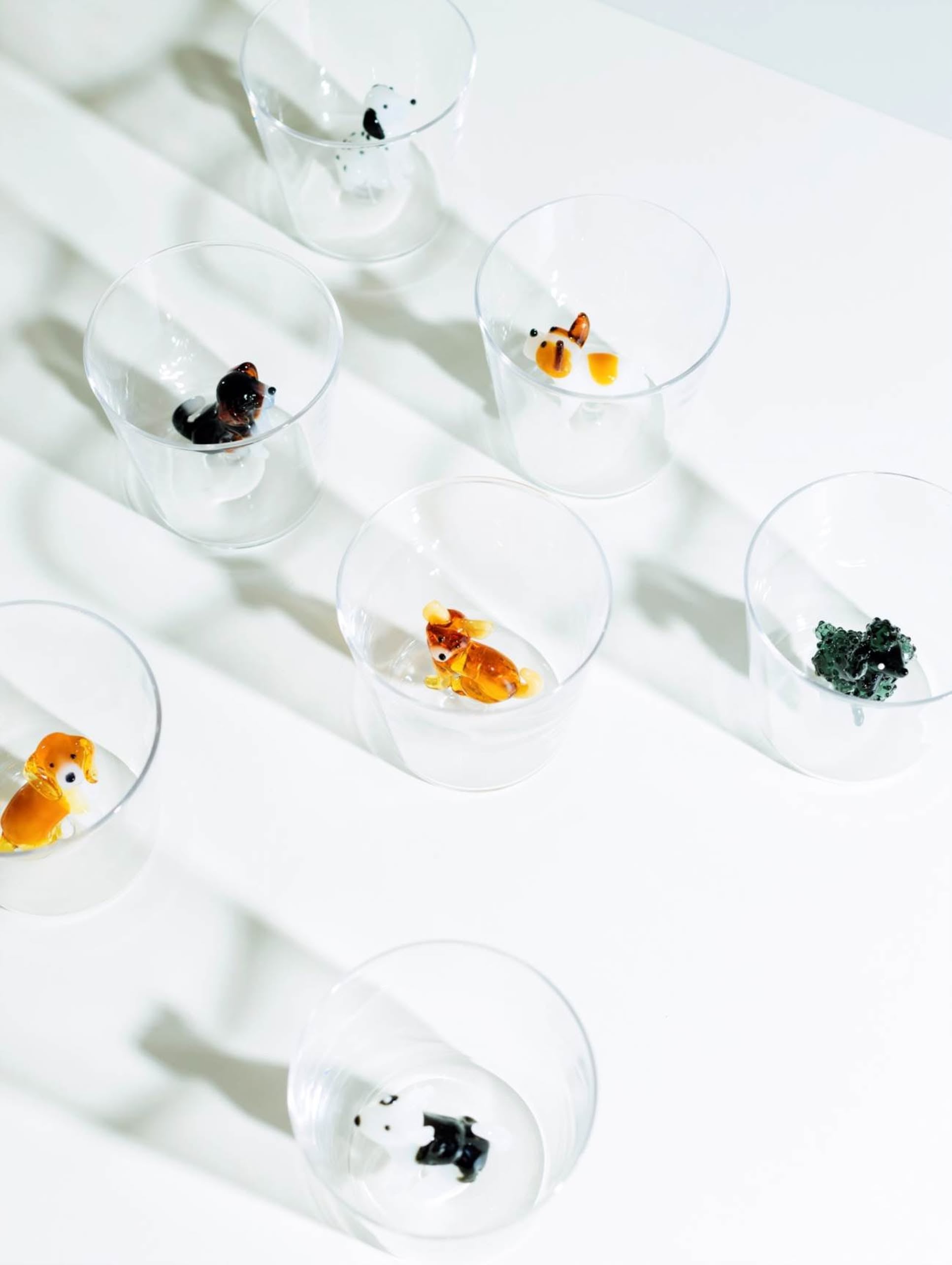 The Ichendorf Milano Dogs Tumbler – English Foxhound is a handmade glass by Ichendorf Milano, shown on a white surface with a small English Foxhound figurine at the bottom. The bright lighting creates soft shadows around the tumbler.