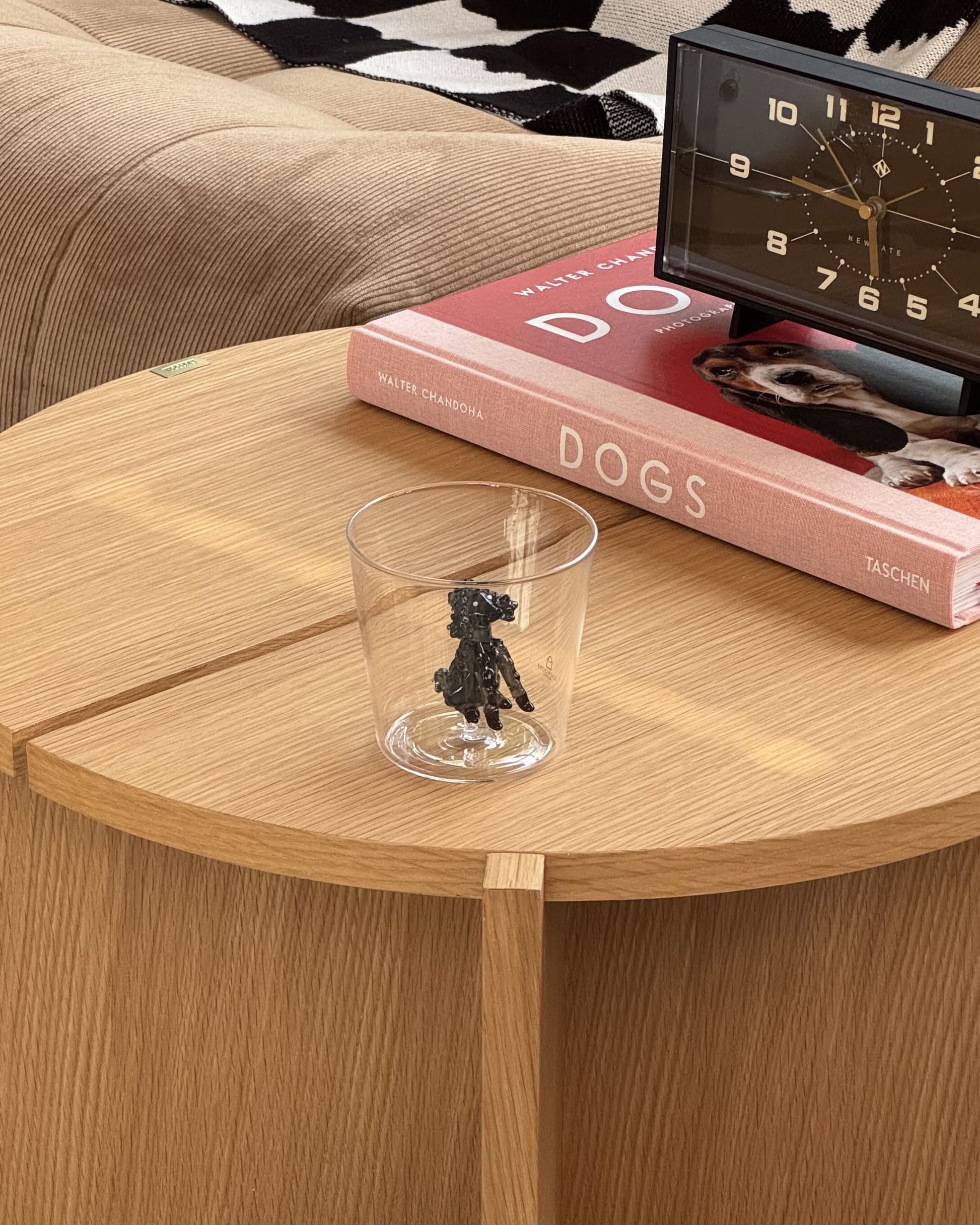 The Ichendorf Milano Dogs Tumbler - Poodle sits on a round wooden table beside a red DOGS book and black clock, with a beige couch draped in a black-and-white blanket in the background.