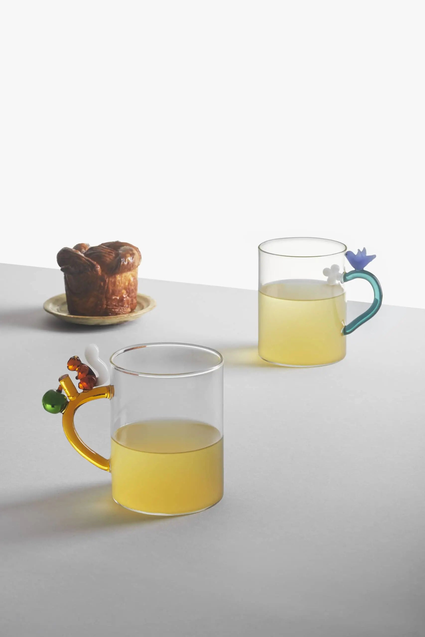 Two Ichendorf Milano Fruits and Flower Mugs, filled with yellow liquid, sit on a white surface beside a small pastry in a bright, minimalist setting.