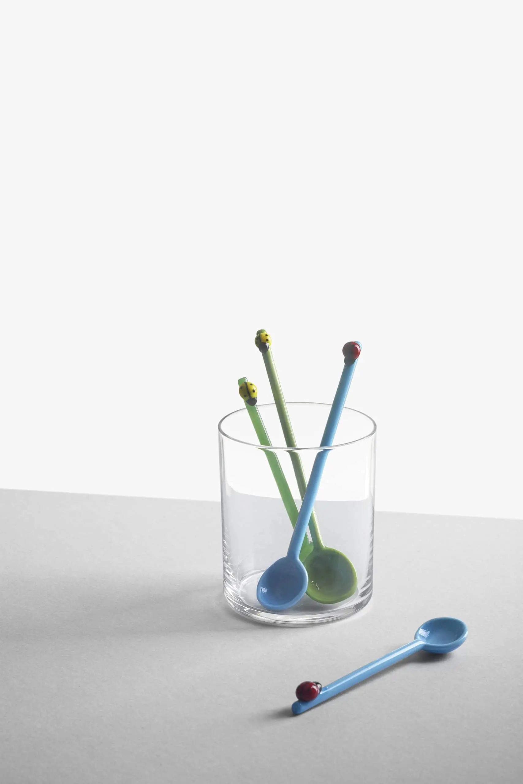 A clear glass holds two green and one blue Ichendorf Milano Fruits and Flower Teaspoons; another blue spoon rests on the gray surface. The background is plain white and light gray.