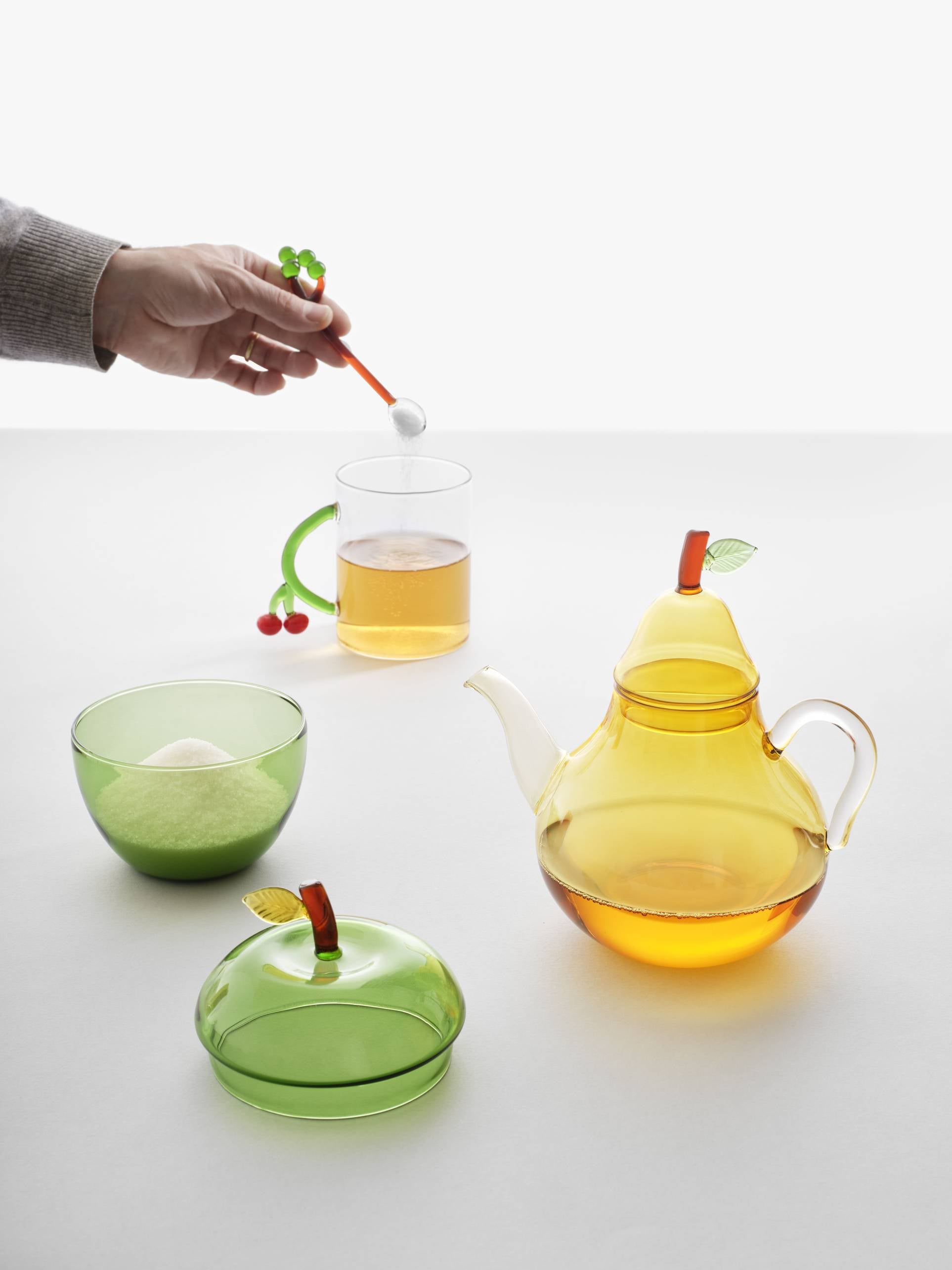 A hand adds a sugar cube to a glass of tea using red cherry-shaped tongs from Ichendorf Milano’s Fruits and Flowers collection. On the table are a yellow teapot, the Ichendorf Milano Fruits and Flowers Sugar Bowl, and green fruit-style dishes.