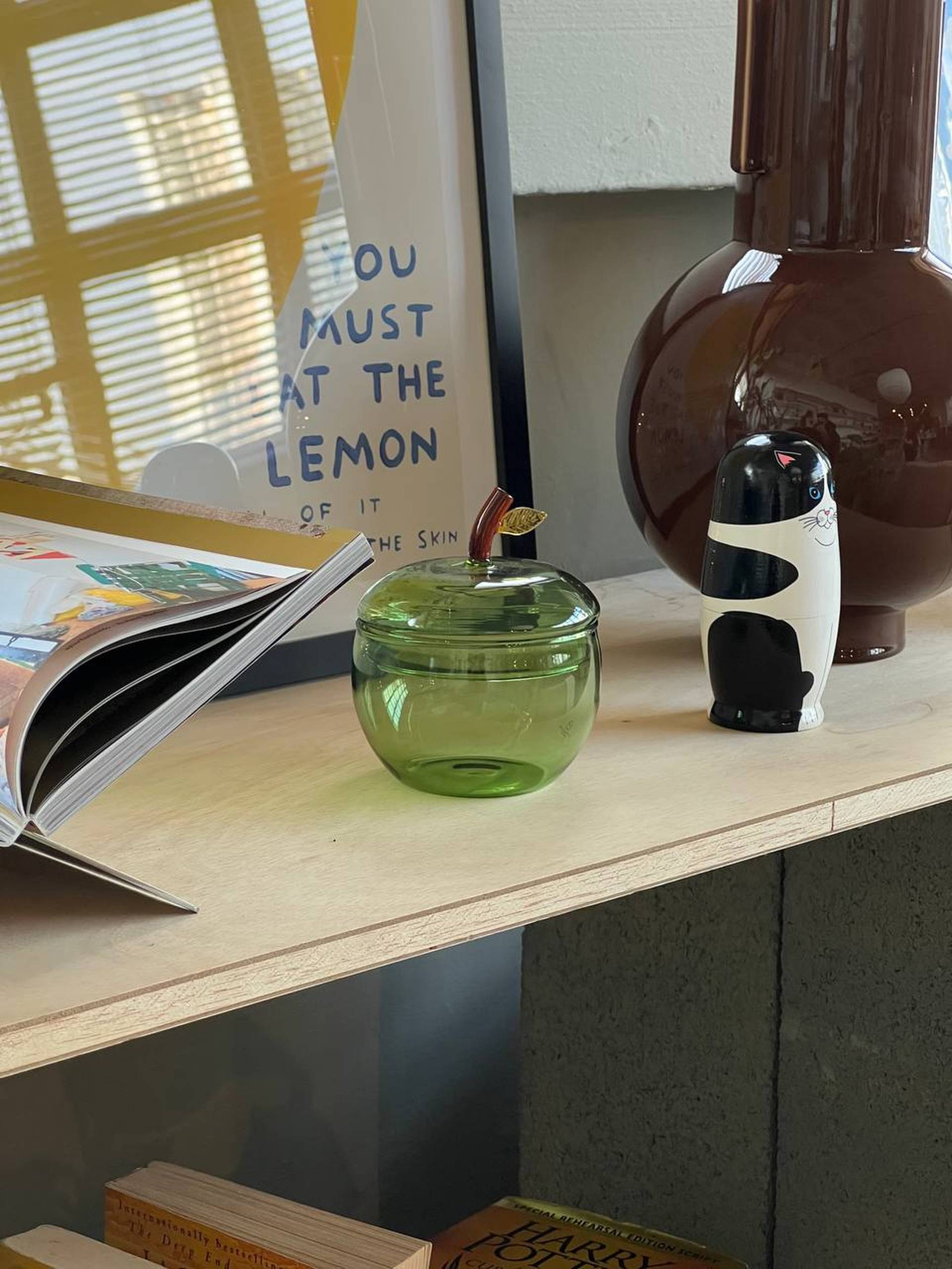 An Ichendorf Milano Fruits and Flowers Sugar Bowl rests on a wooden shelf beside an open book, a brown vase, assorted fruits, and framed art with the phrase YOU MUST AT THE LEMON.