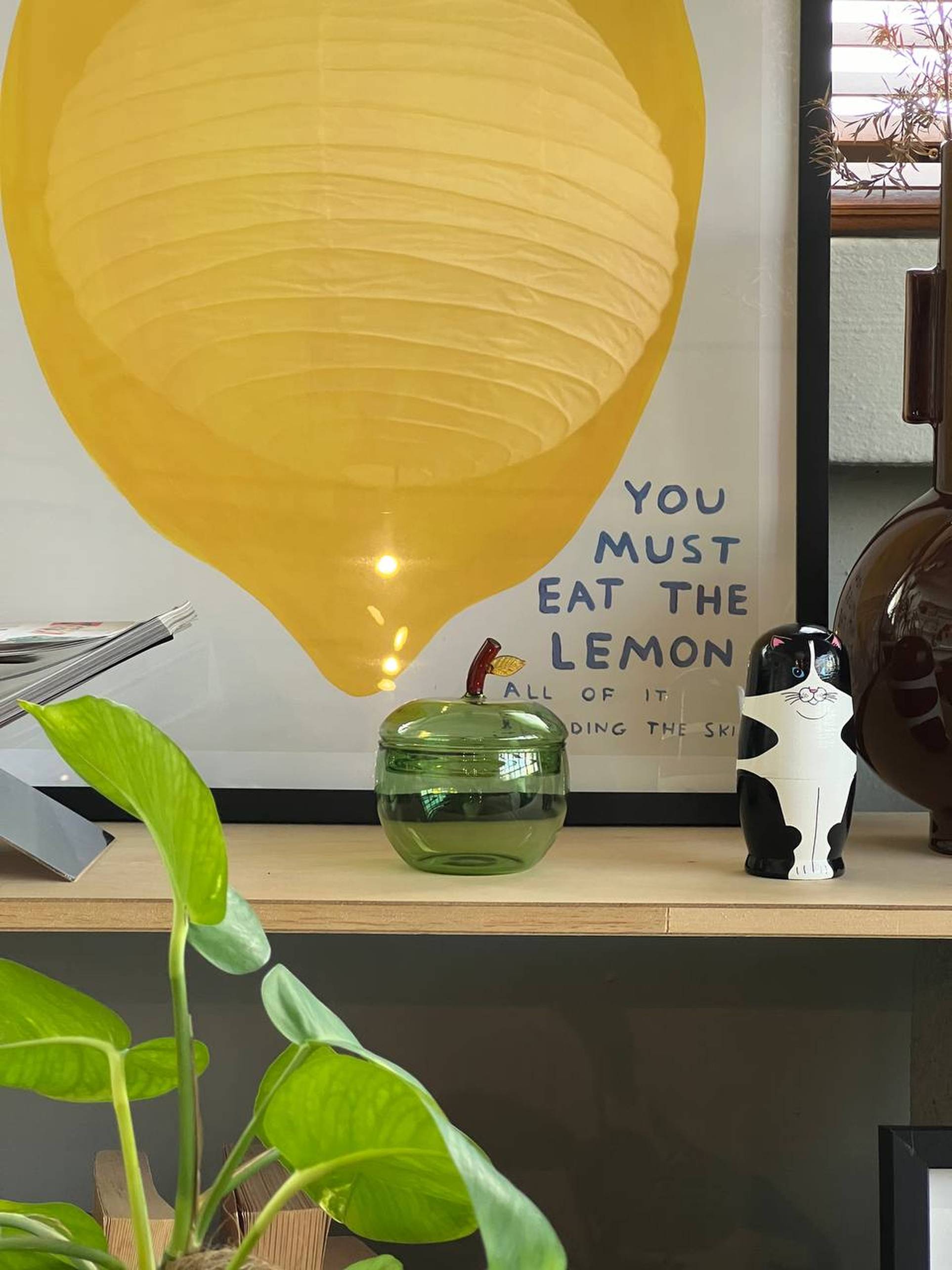 A shelf displays green plants, the Ichendorf Milano Fruits and Flowers Sugar Bowl, a green glass jar, a black and white cat figurine, and a framed poster of a large yellow lemon with the text YOU MUST EAT THE LEMON ALL OF IT.