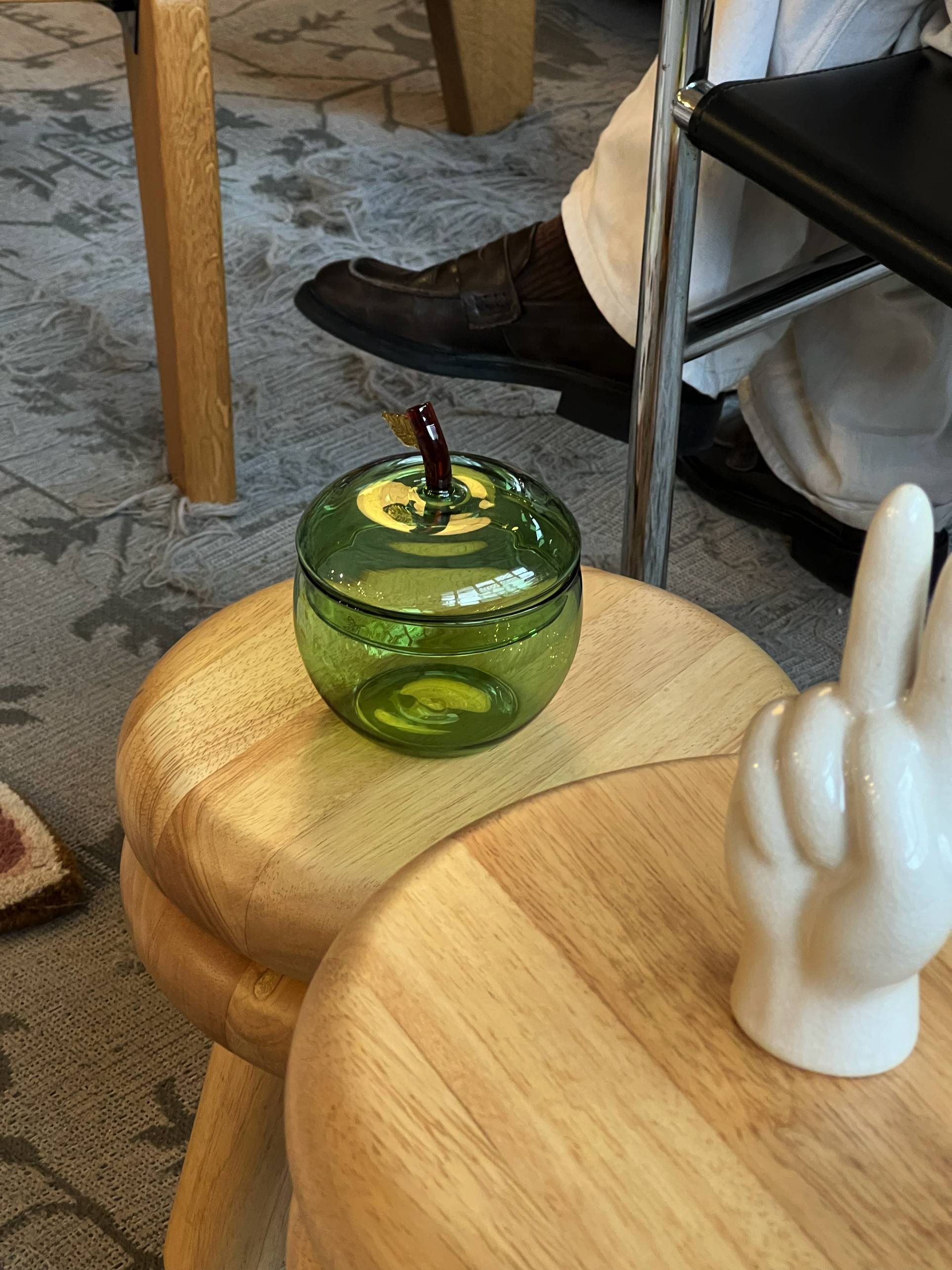 An Ichendorf Milano Fruits and Flowers Sugar Bowl shaped like a green apple sits on a wooden table next to a white ceramic hand making a peace sign, with colorful fruits and someone in white pants in the background.