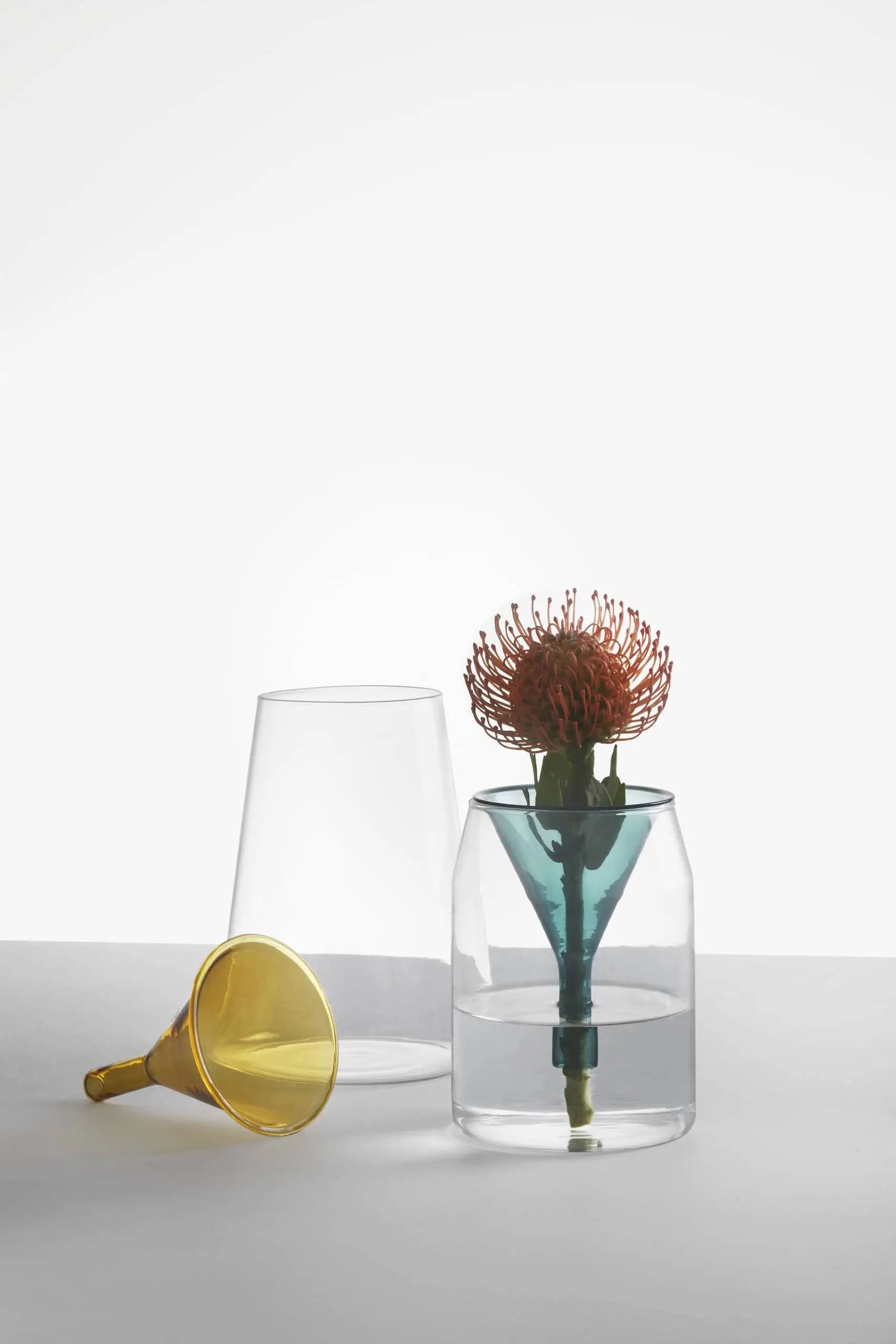 An Ichendorf Milano Kado Single Flower Vase 20cm, hand-flamed glass with water, holds a blue funnel and an orange protea. Beside it, a yellow funnel and an empty glass rest on a white surface against a plain background.