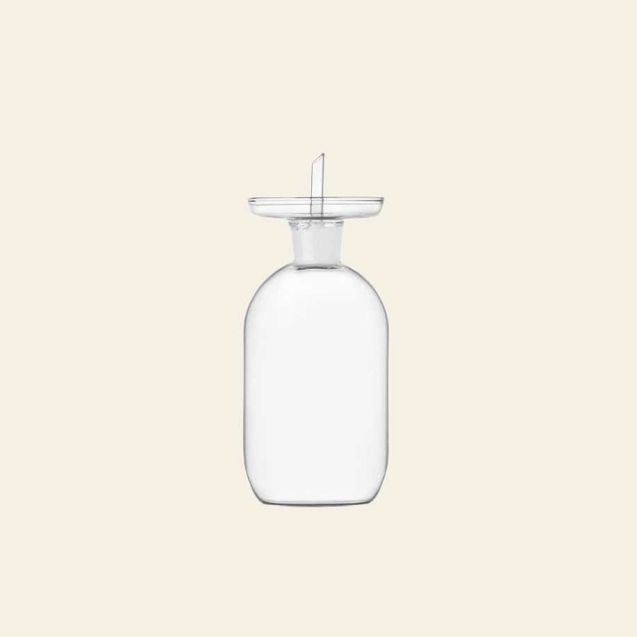 The Ichendorf Milano Lotus Oil Cruet Smooth Medium, a clear glass vessel with a wide neck and flat round stopper, stands upright against a light beige background—ideal for elevating any kitchen space.