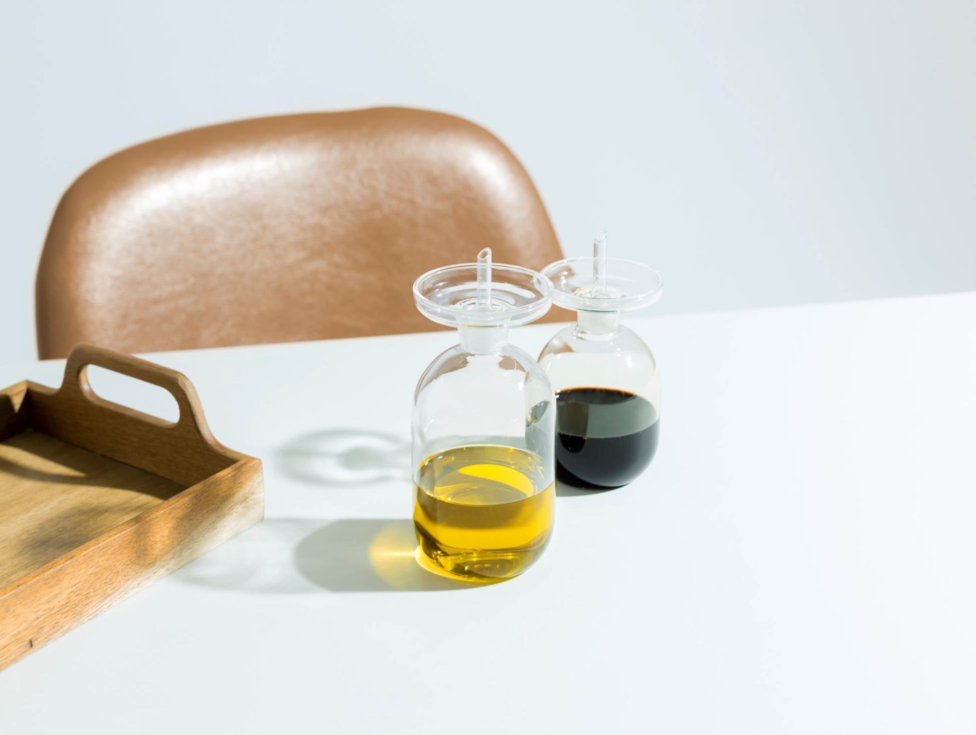 The Ichendorf Milano Lotus Oil Cruet Smooth Medium by Ichendorf Milano, filled with yellow and dark liquids, sits on a white table next to a wooden tray, bringing elegance to the kitchen with a brown chair in the background.