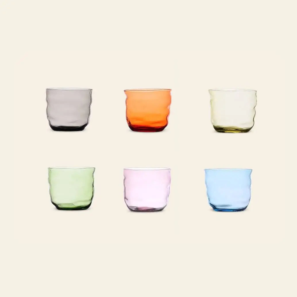 The Ichendorf Milano Poseidon Tumbler Set of 6 features organically shaped, colorful glasses—gray, orange, yellow, green, pink, and blue—arranged in two rows of three on a plain light background.
