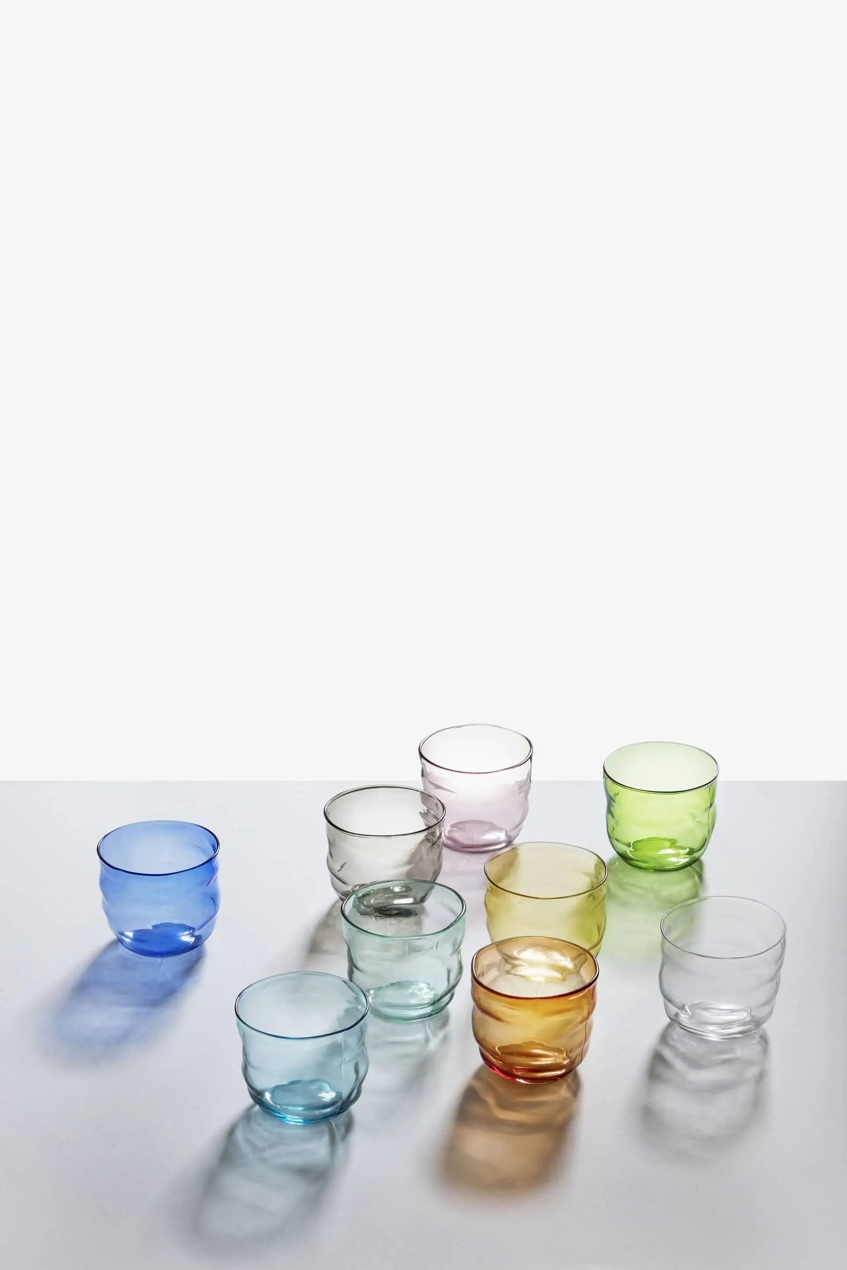 Six Ichendorf Milano Poseidon Tumblers in blue, green, yellow, amber, pink, and clear—each with organic shapes—are arranged on a reflective white surface against a simple light gray backdrop.