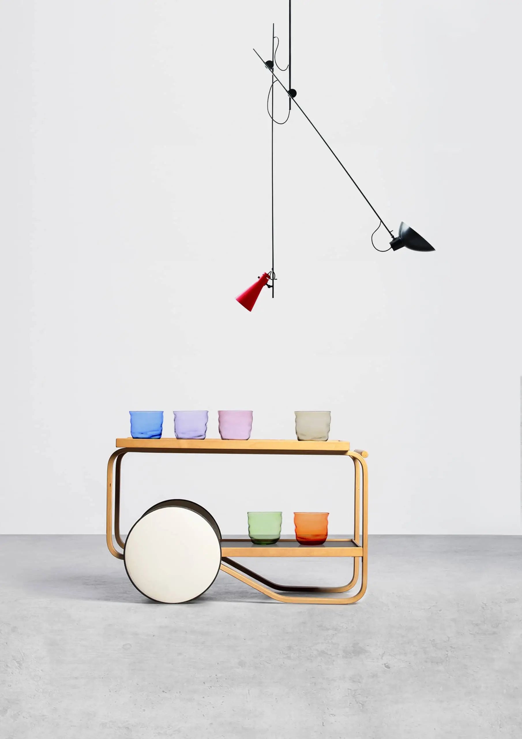 A minimalist scene features a wooden bar cart displaying vibrant Ichendorf Milano Poseidon Tumblers on both shelves, a large circular accent on one side, and two modern pendant lights—one red, one black—hanging overhead.