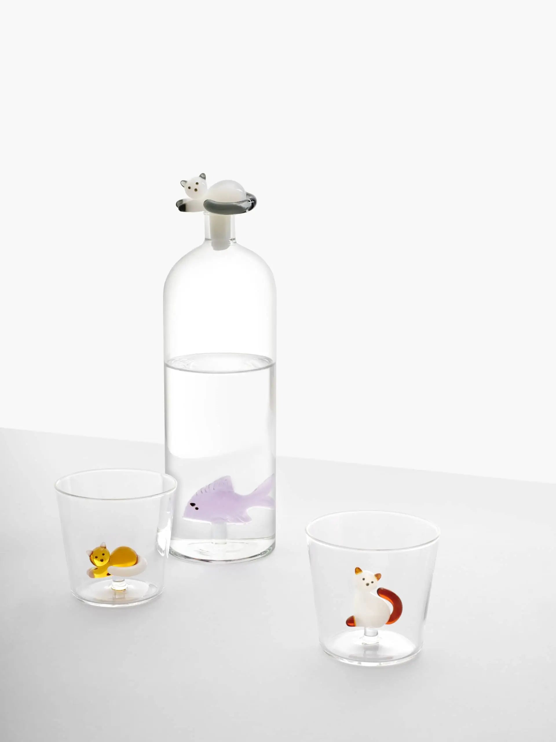 The Ichendorf Milano Tabby Cat Bottle by Ichendorf Milano is a clear glass bottle with water, featuring a white cat figurine on the lid and a purple fish inside. Nearby, two handcrafted glasses each hold an adorable yellow squirrel and red fox figure.