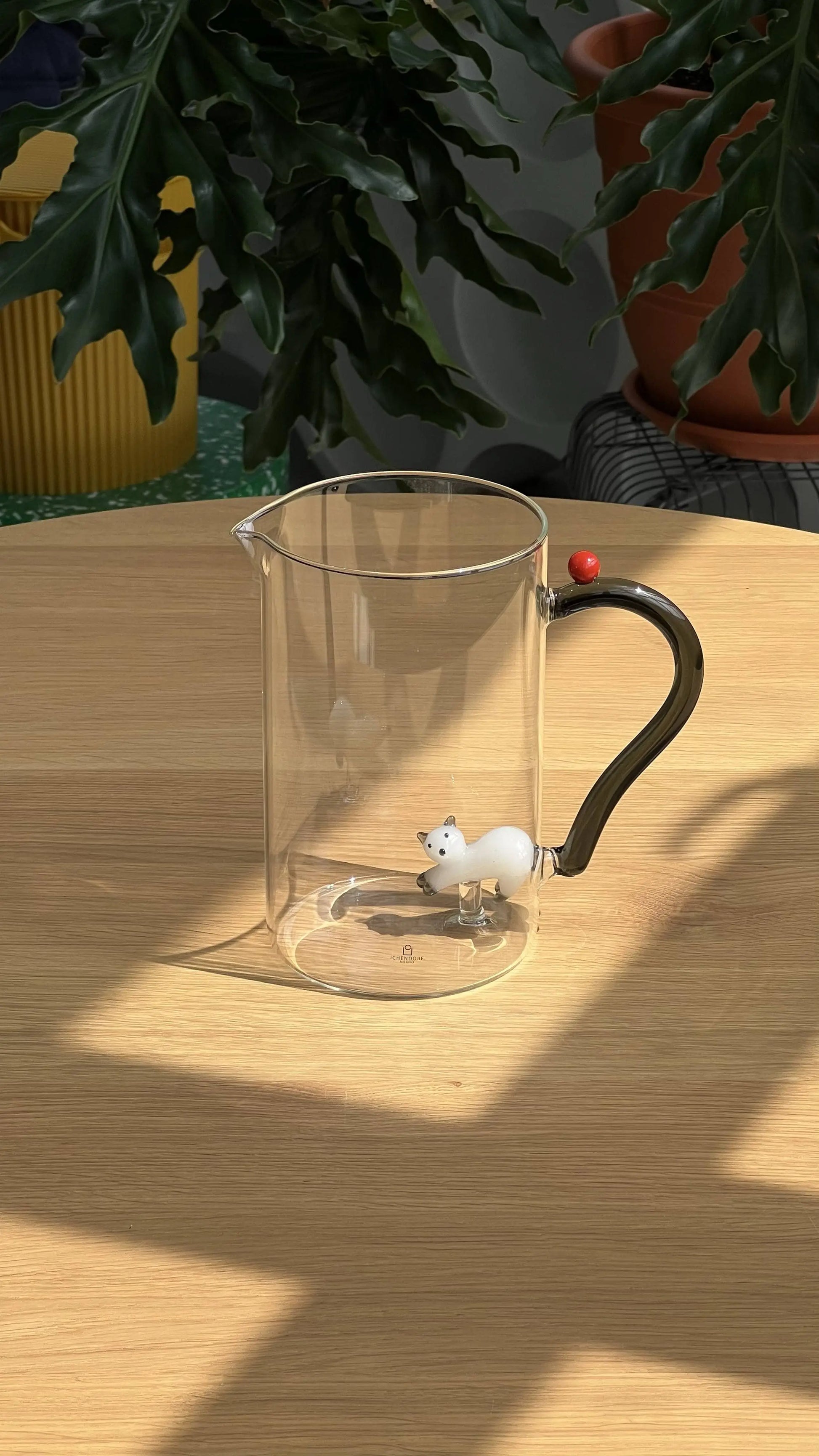 The Ichendorf Milano Tabby Cat Jug, a clear glass handcrafted piece with a black handle by Ichendorf Milano, sits on a wooden table. A white cat figurine rests inside the jug, with green potted plants in the background.