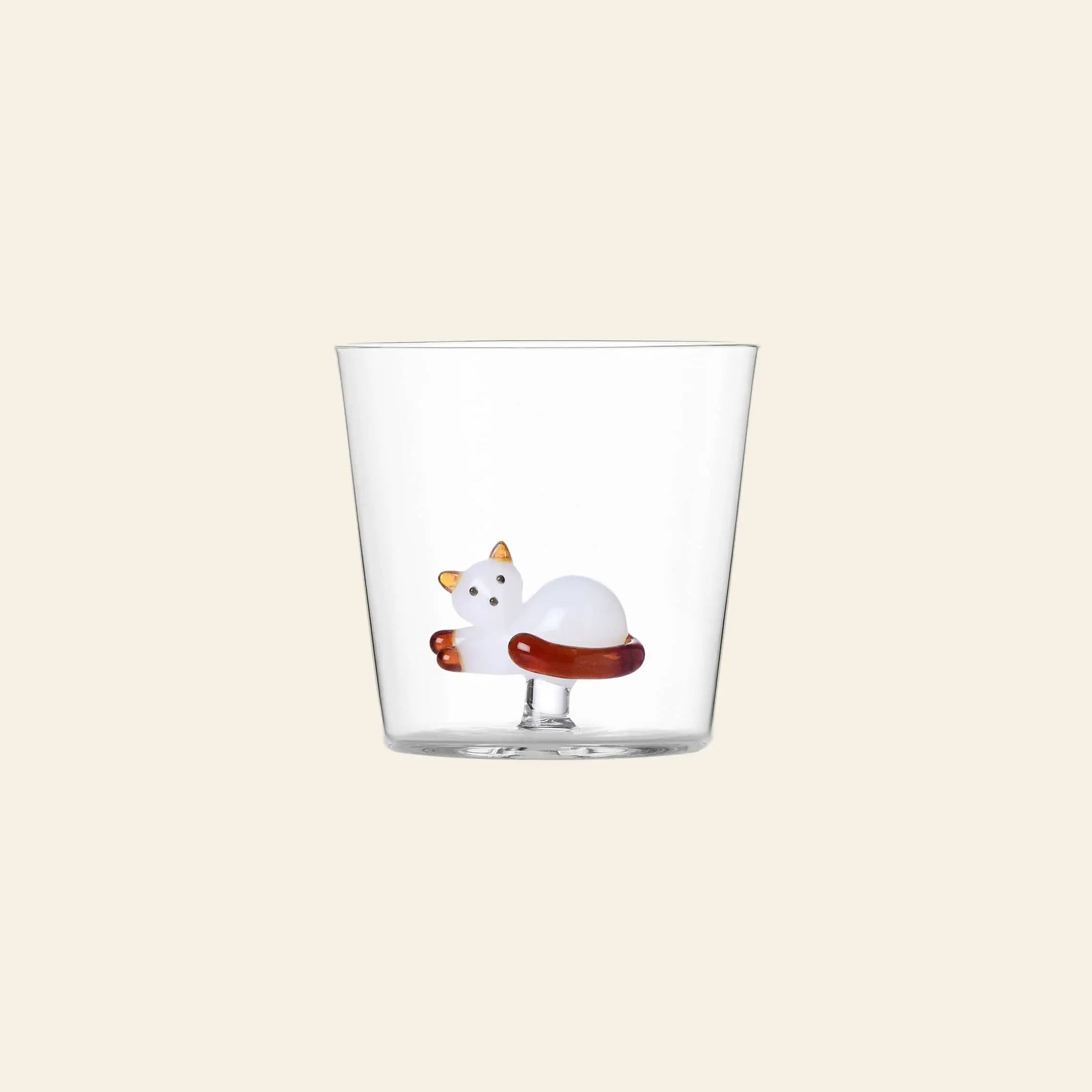 The Ichendorf Milano Tabby Cat Tumbler by Ichendorf Milano is a clear glass featuring a sculpted white cat with orange ears and tail at the bottom, set against a plain light beige background for a charming touch.