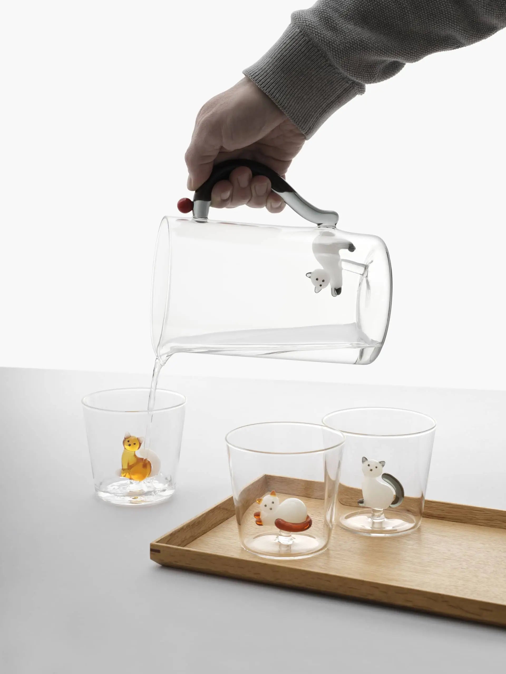 A hand pours water from an Ichendorf Milano pitcher into one of three Tabby Cat Tumblers by Ichendorf Milano, each featuring a duck, cat, or dog figurine at the bottom. The pitcher handle is adorned with a polar bear figurine.
