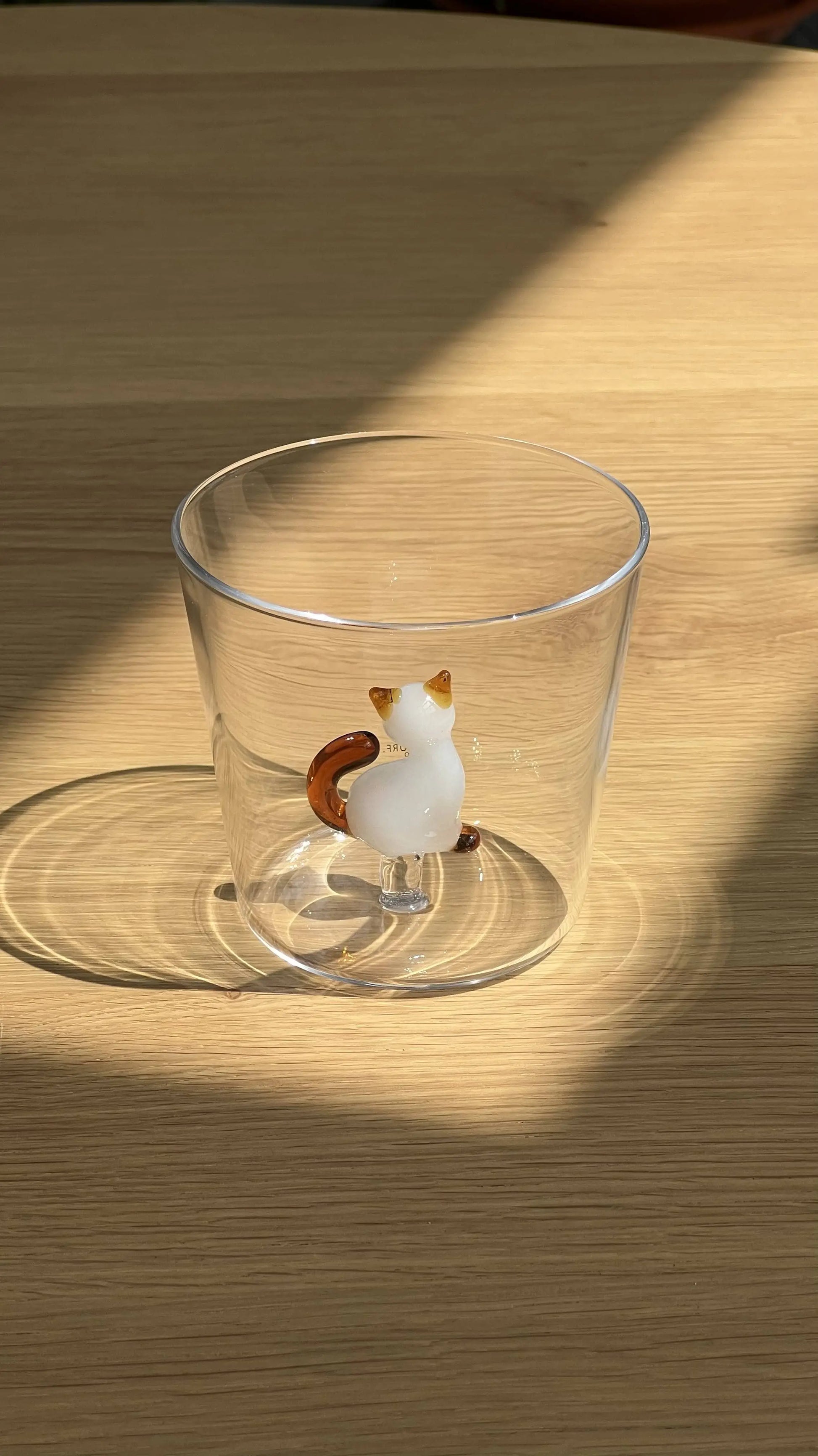 The Ichendorf Milano Tabby Cat Tumbler by Ichendorf Milano sits on a wooden surface, sunlight casting shadows. Inside, a small cat figurine from the Tabby Cat collection is attached to the base—perfect for feline fans.