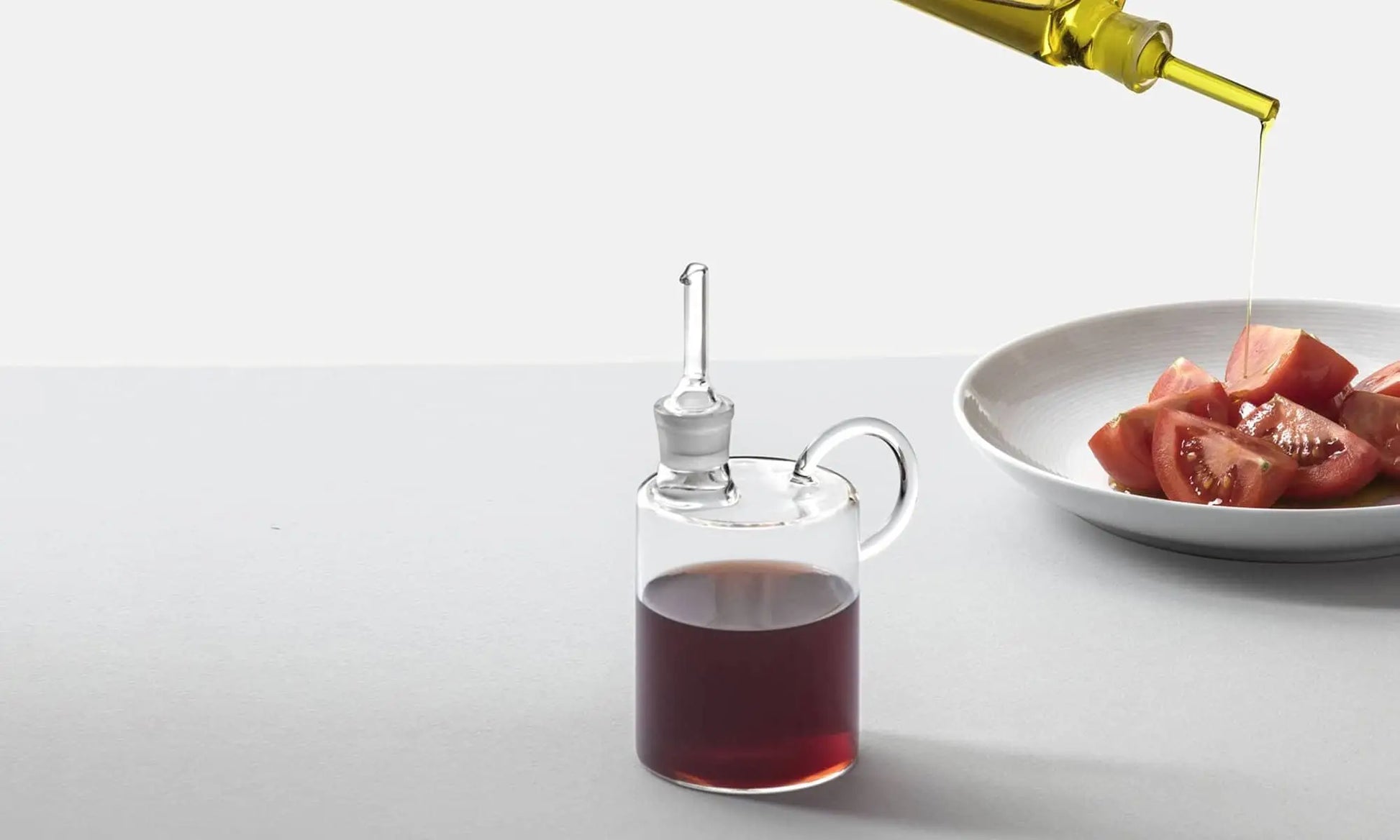 The Ichendorf Milano Tokio Balsamic Vinegar Bottle by Ichendorf Milano rests on a light surface, while olive oil is poured over tomato wedges in the background, showcasing this elegant culinary accessory.