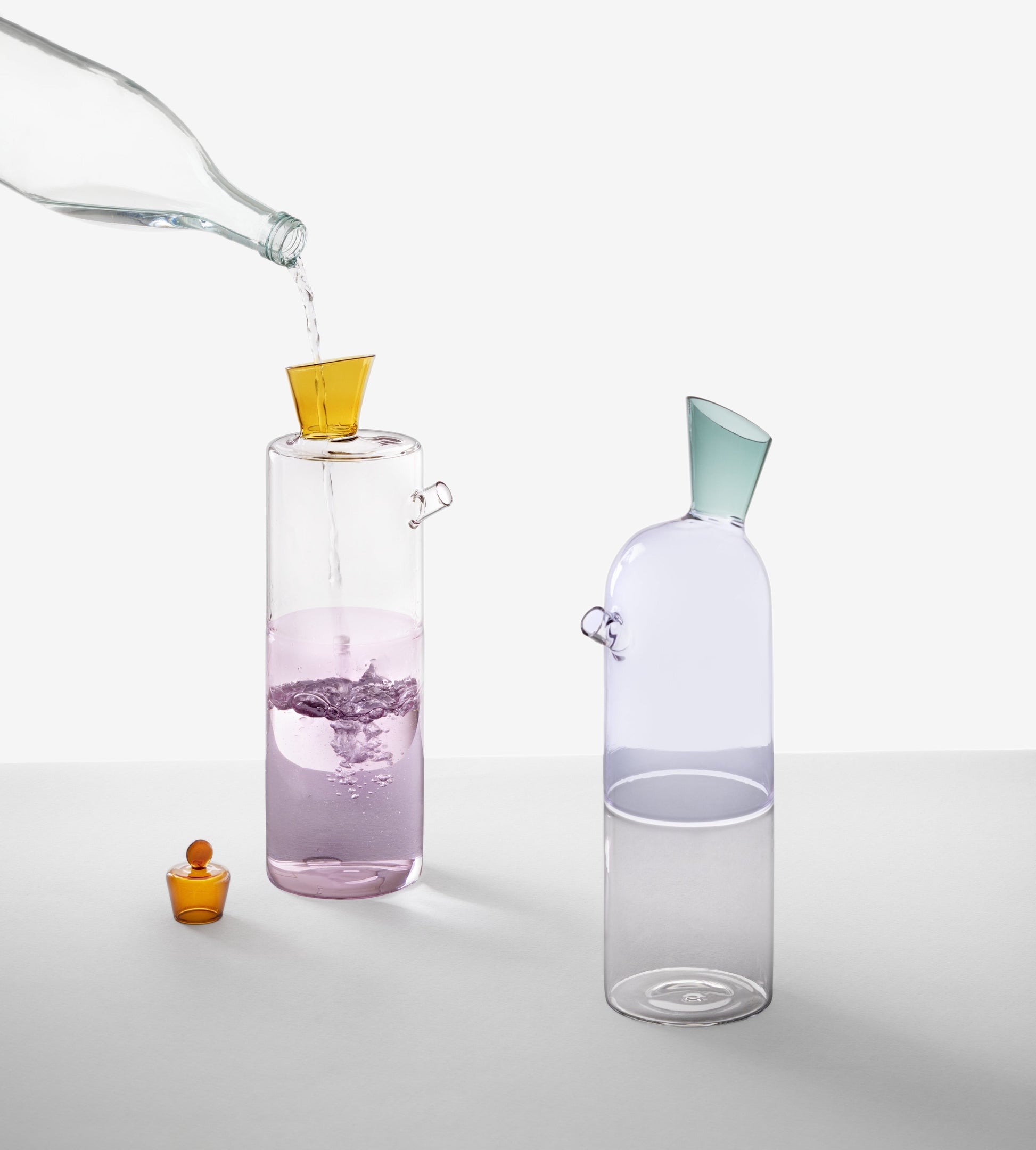 A hand pours water from the Ichendorf Milano Travasi Bottle by Ichendorf Milano into a tall glass with a yellow funnel, highlighting geometric design. Another glass with a blue funnel and an orange stopper are seen beside it.