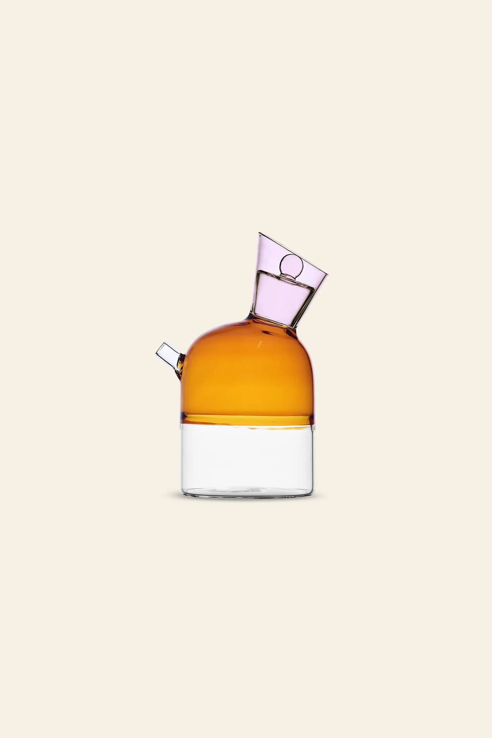 The Ichendorf Milano Travasi Oil Bottle by Ichendorf Milano features a modern glass design with an amber top, clear base, side spout, and tilted neck—an elegant geometric piece ideal for stylish table settings, shown on a plain beige background.