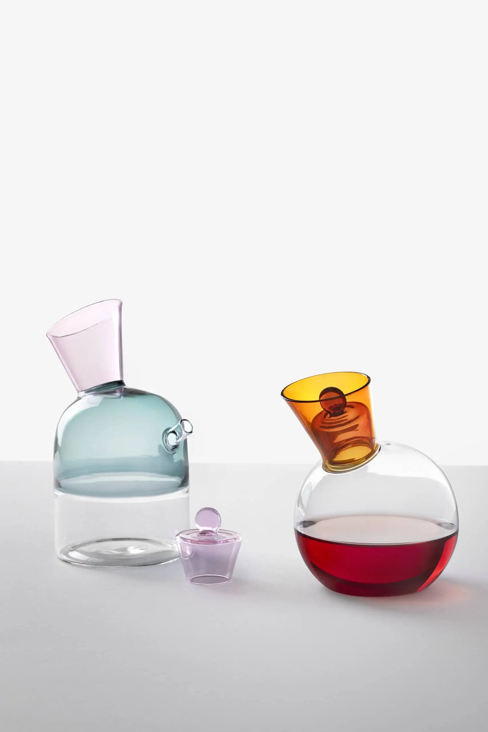Three glass vessels in modern, geometric styles: one light blue with a slanted neck, one small purple with round stopper, and Ichendorf Milano Travasi Oil Bottle—round, amber neck, red liquid—perfect for contemporary table settings.